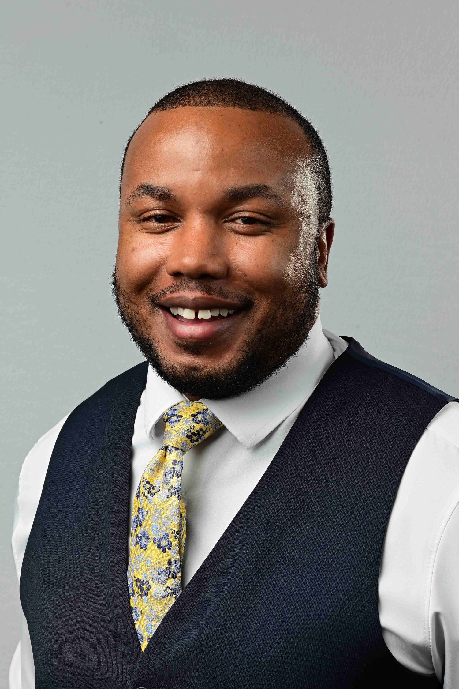Smiling professional man in navy vest and floral yellow tie poses for a business headshot on gray background.
