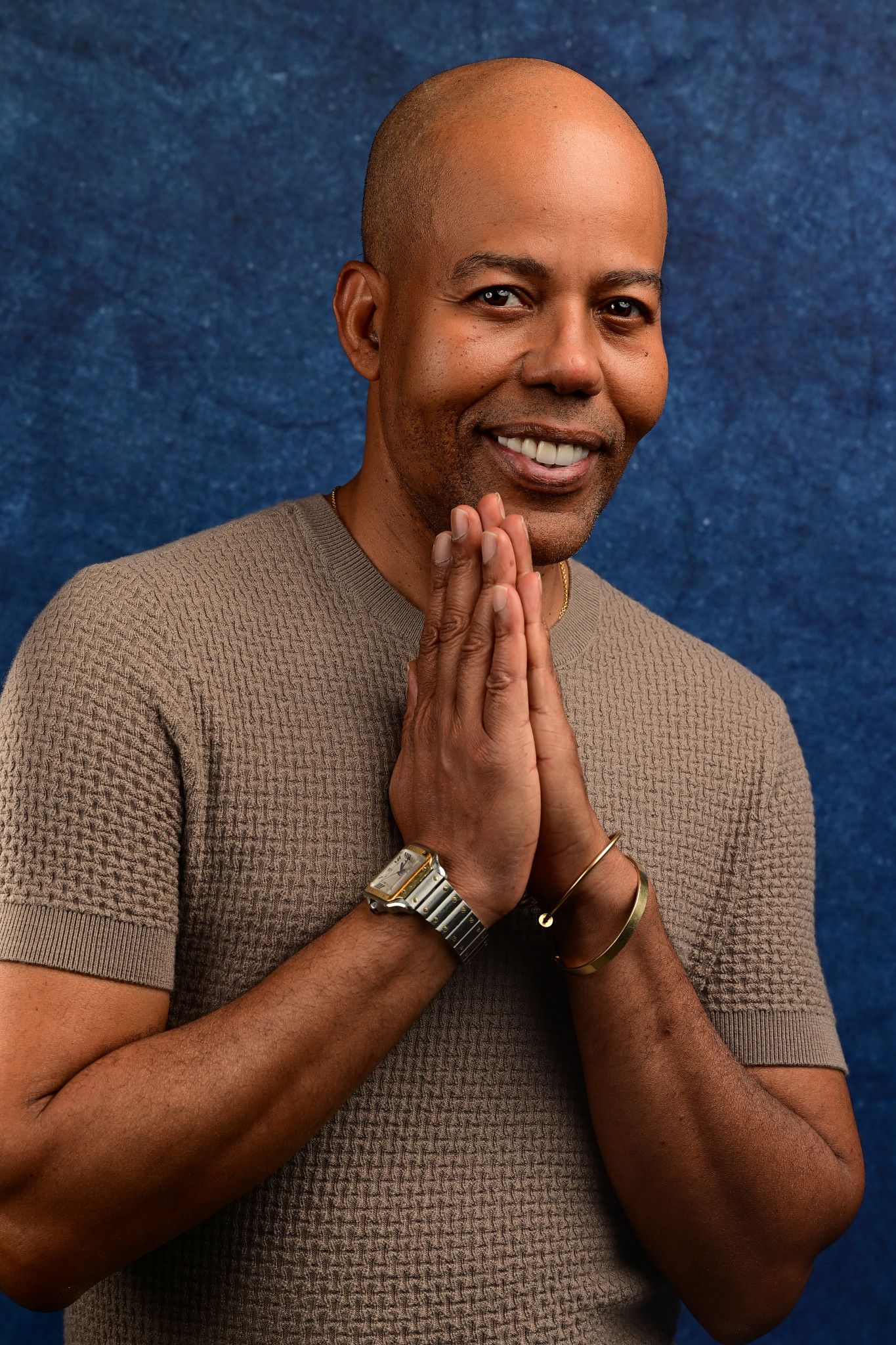 Smiling bald man in a textured taupe knit shirt presses his palms together in front of his chin against a deep blue backdrop.