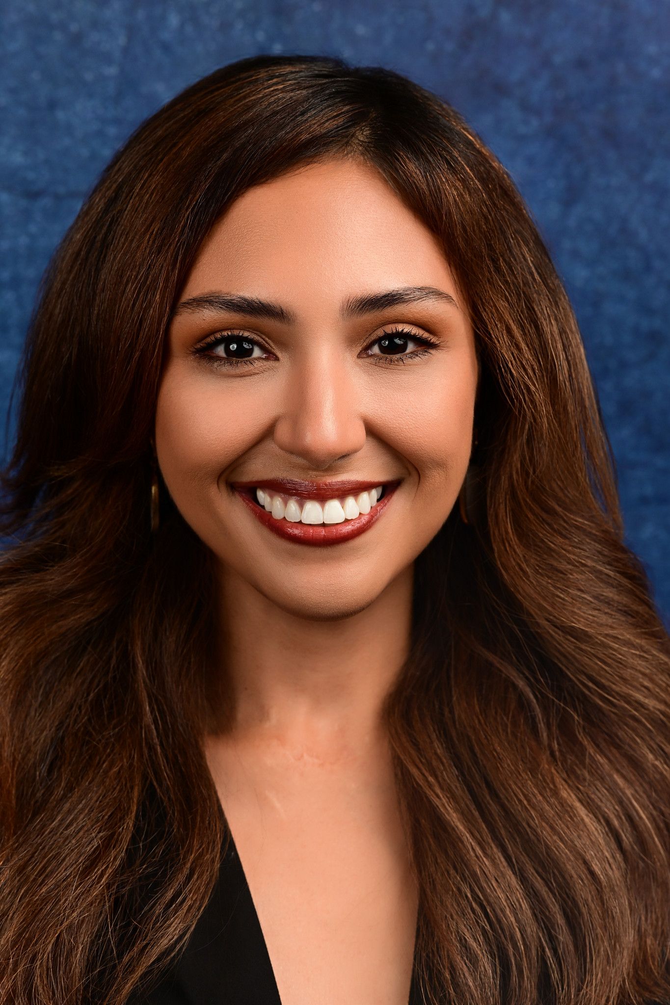 Smiling woman with long wavy brown hair and red lipstick wearing a black top in a professional headshot against a blue textured background.