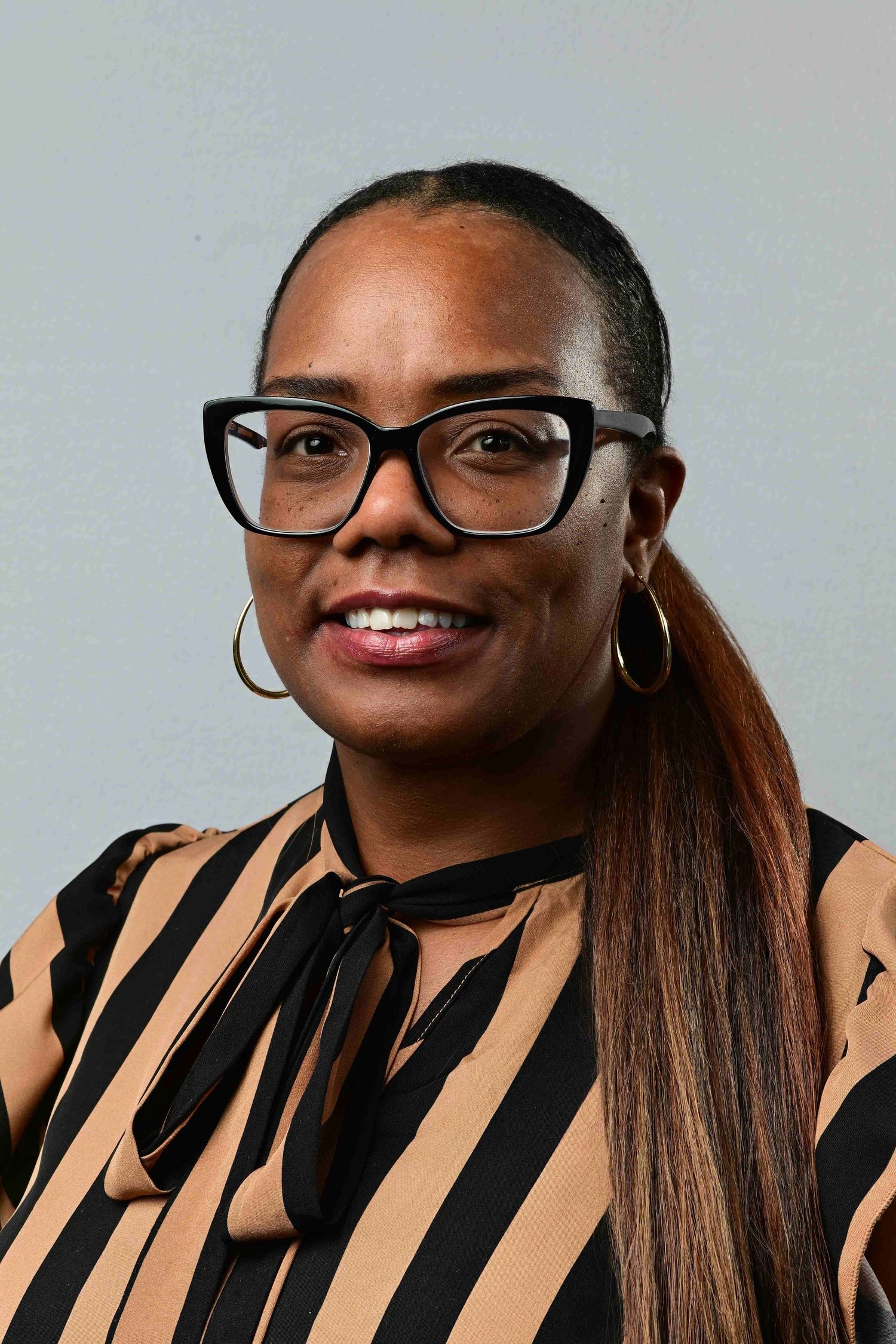 Professional headshot of a woman wearing black-framed glasses, hoop earrings, and a striped black and tan blouse.