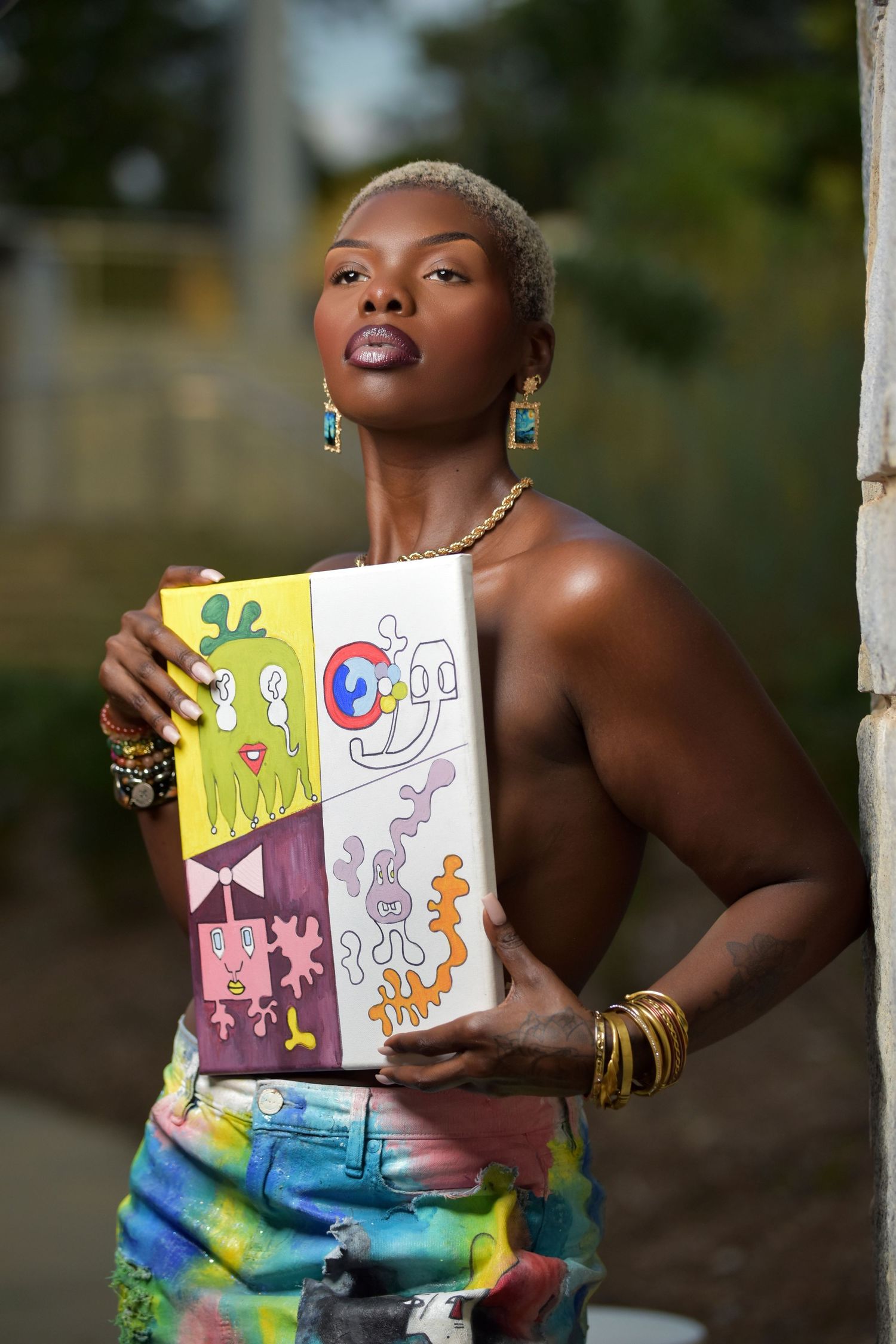 Black woman artist holding colorful abstract painting canvas outdoors, wearing tie-dye jeans and gold jewelry.