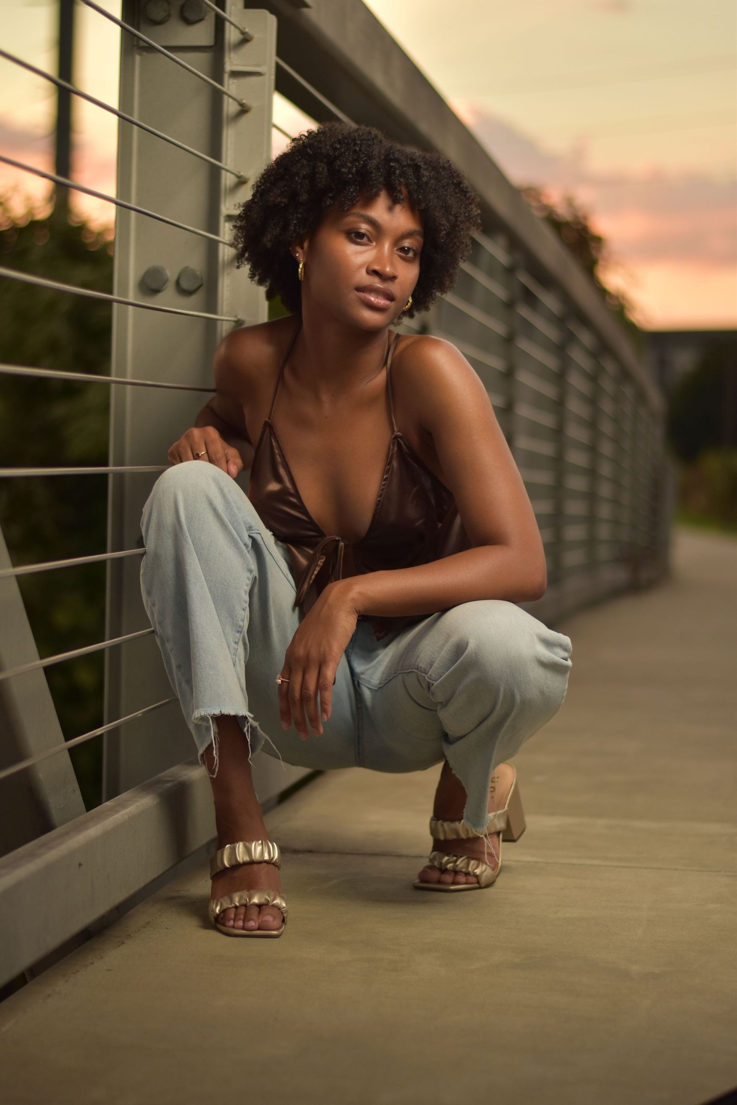 Stylish Black woman in brown halter top and distressed jeans posing on bridge at golden hour.