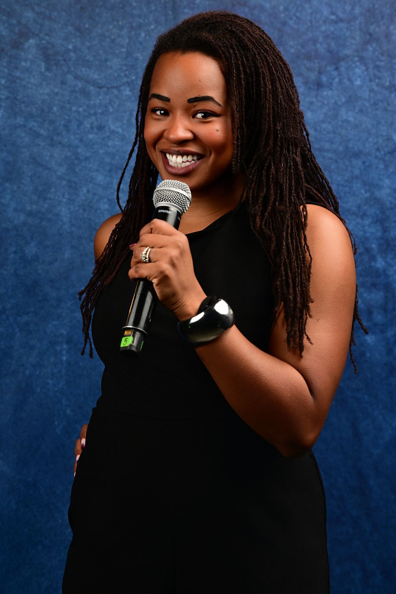Smiling woman with long locs in a black sleeveless dress holds a microphone against a textured blue studio backdrop.