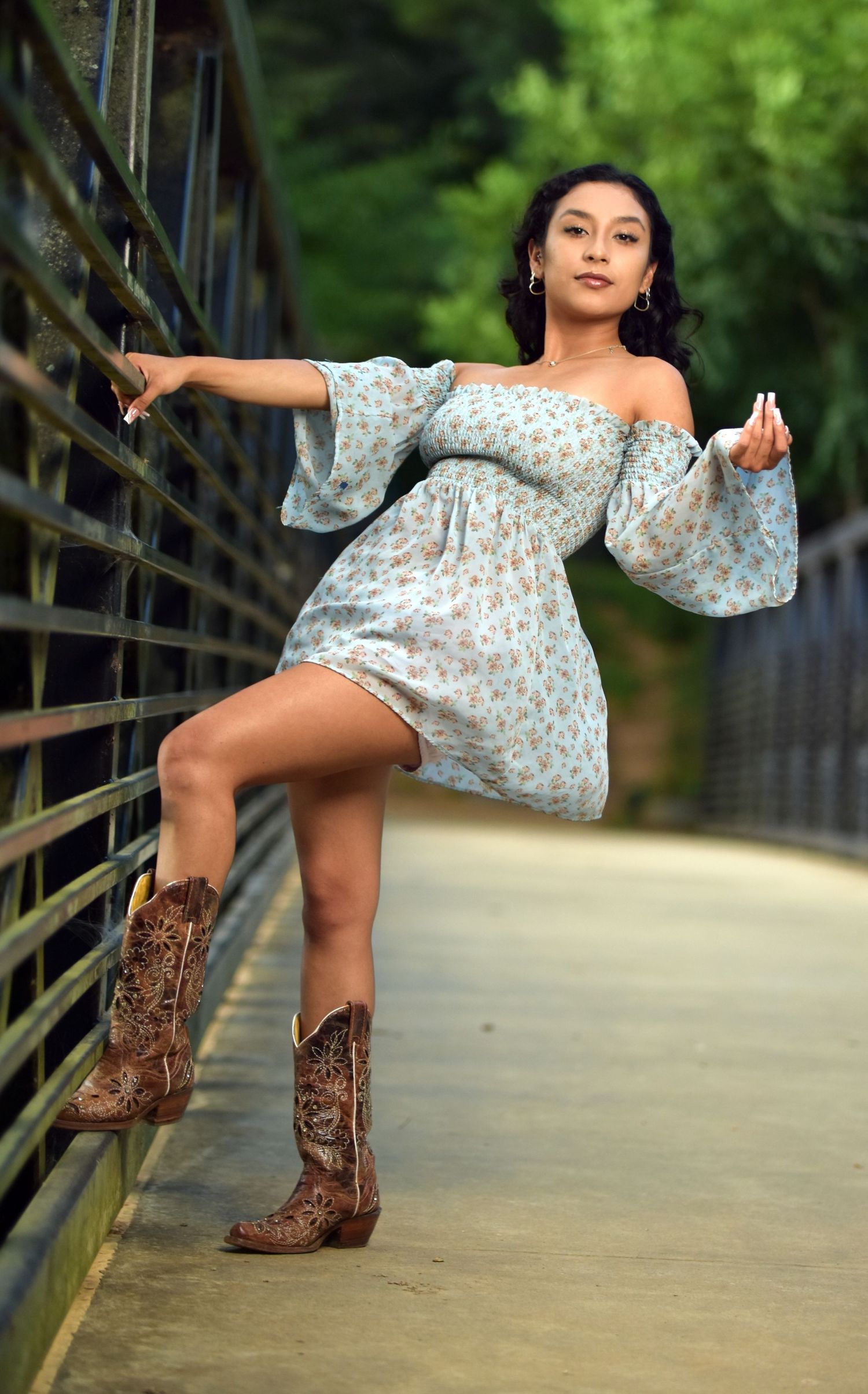 Young woman in floral off-shoulder dress and cowboy boots posing dynamically on a bridge walkway.