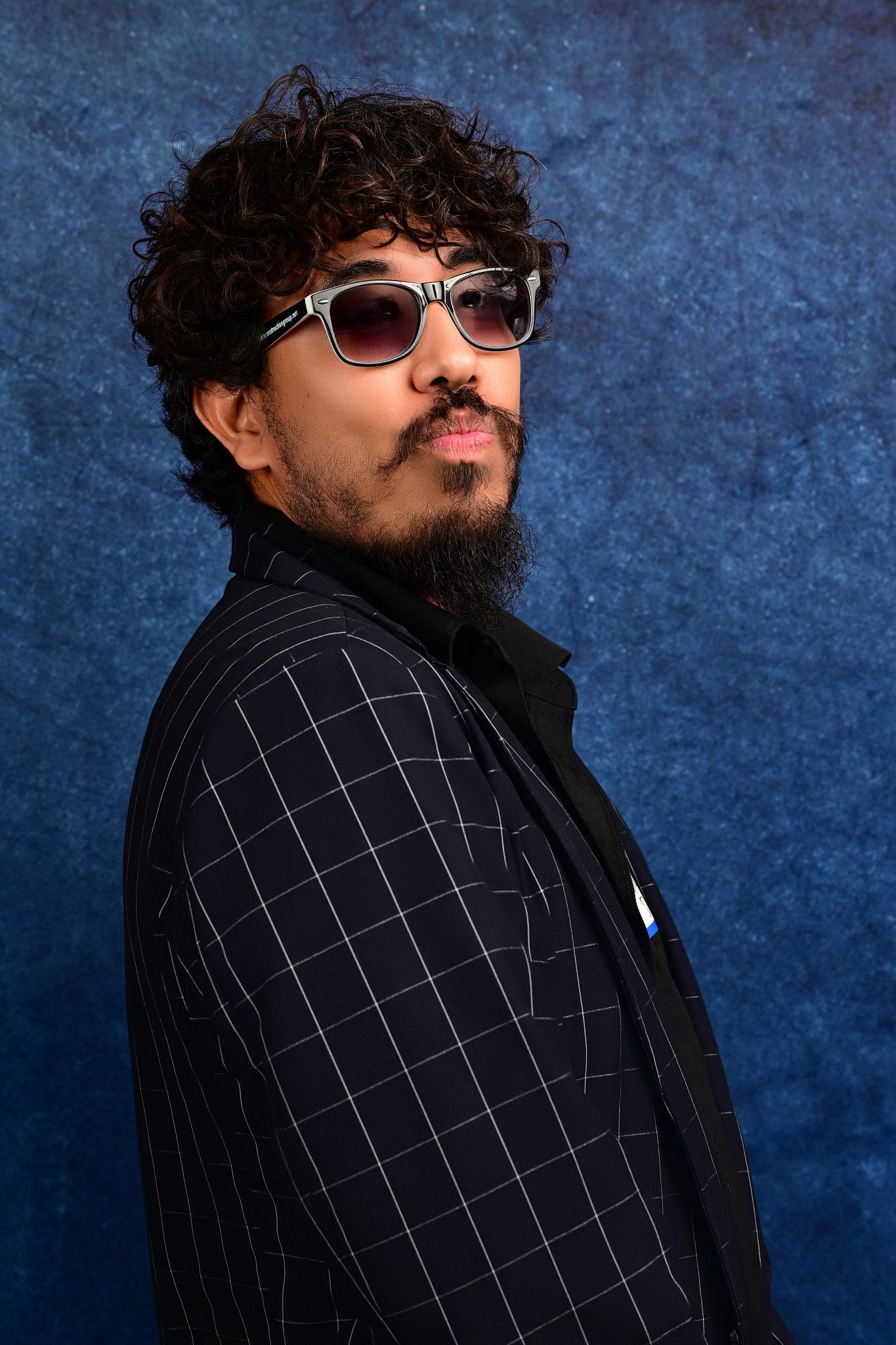 Man with curly dark hair, a beard and tinted sunglasses poses in profile wearing a black windowpane-check blazer against a textured blue bac