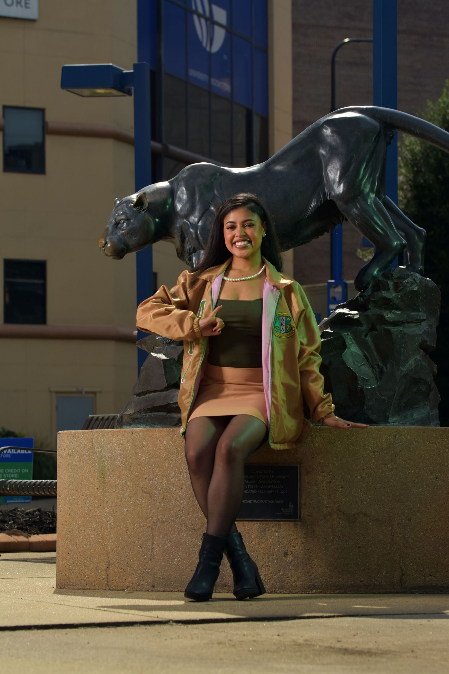Young woman in tan jacket and mini skirt poses smiling beside a bronze panther statue outside a university building.