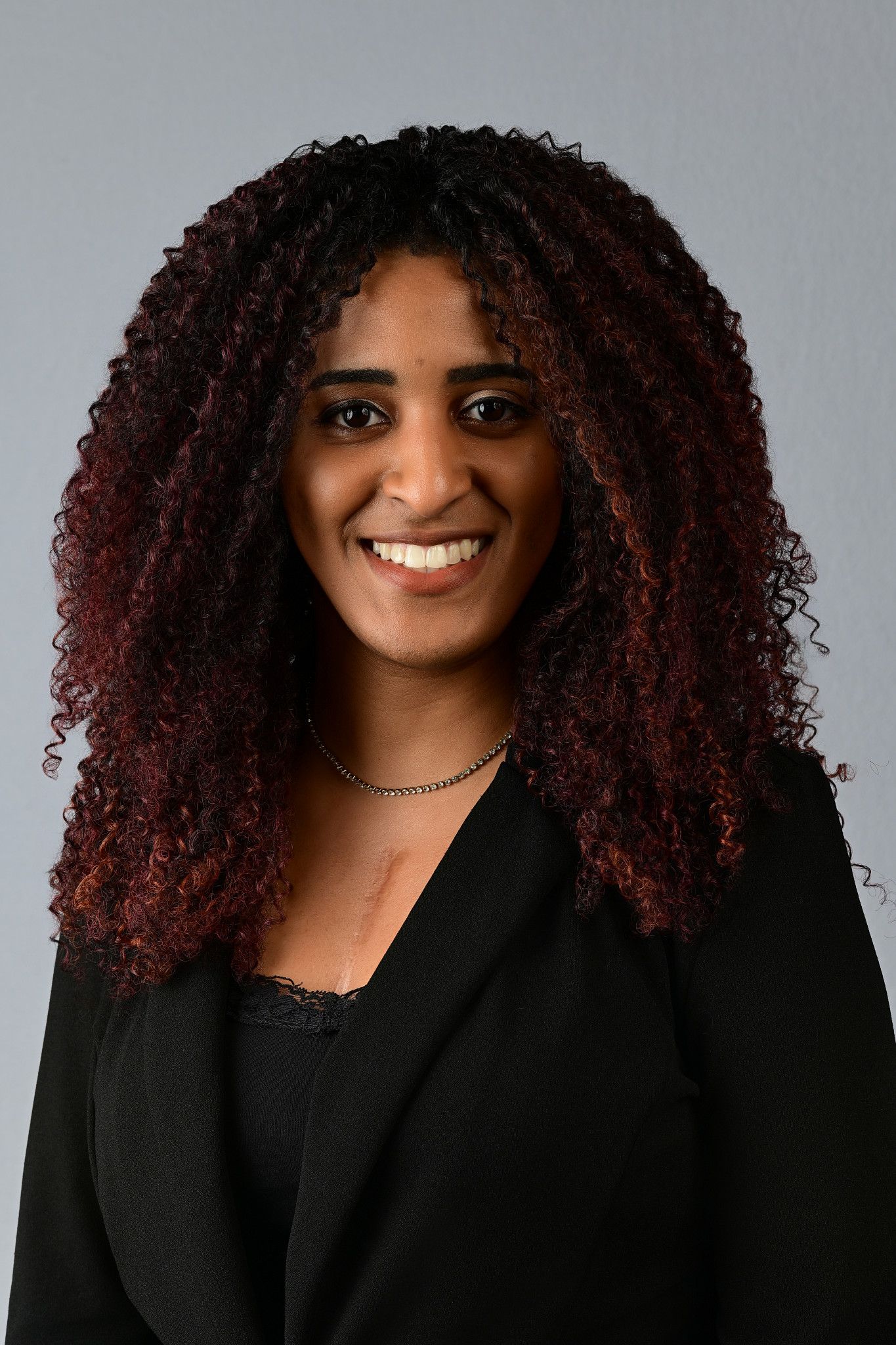 Professional headshot of a smiling woman with curly red-black hair wearing a black blazer against a gray background.