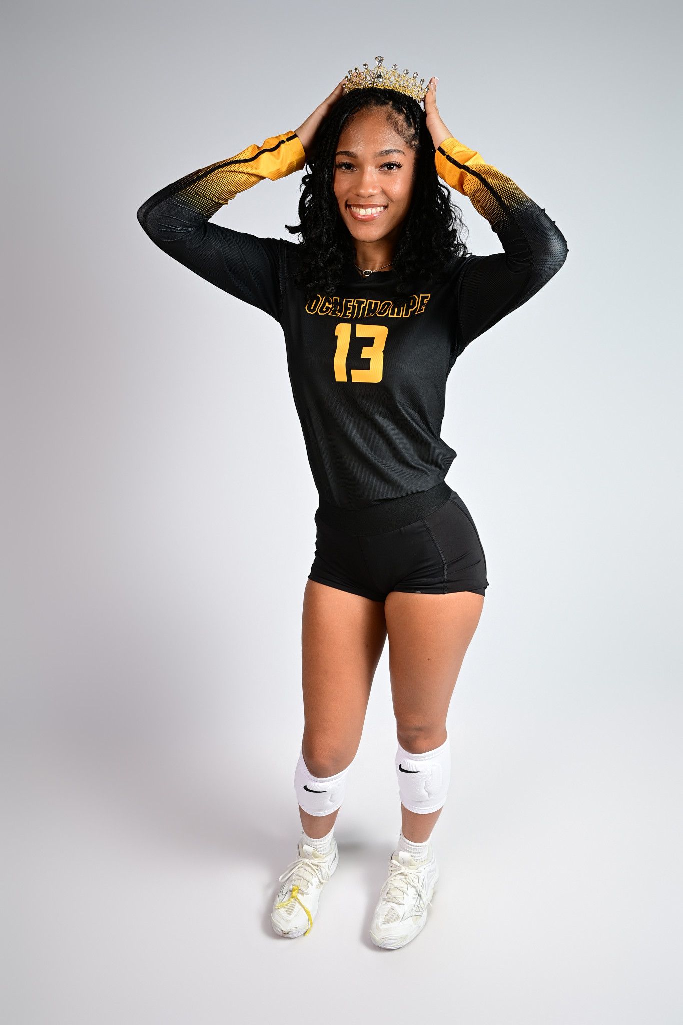 Smiling volleyball player wearing number 13 jersey and crown poses in black and gold uniform with knee-high socks.