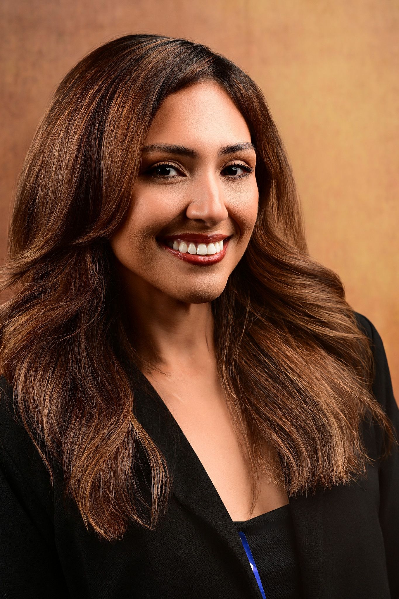 Smiling woman with long wavy brown hair and red lipstick wearing a black blazer in a professional headshot against a warm golden background.