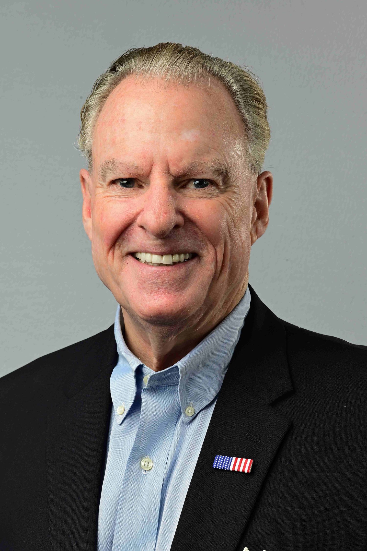 Smiling senior man in black blazer with American flag pin and light blue shirt poses for professional headshot.
