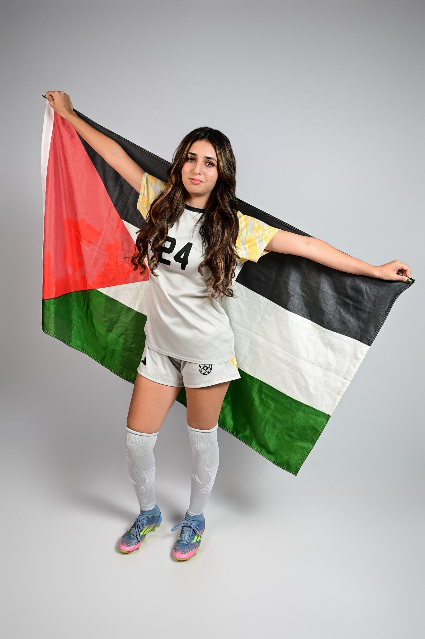 Female soccer player in white uniform #24 holding a Palestinian flag, wearing white knee socks and colorful cleats.