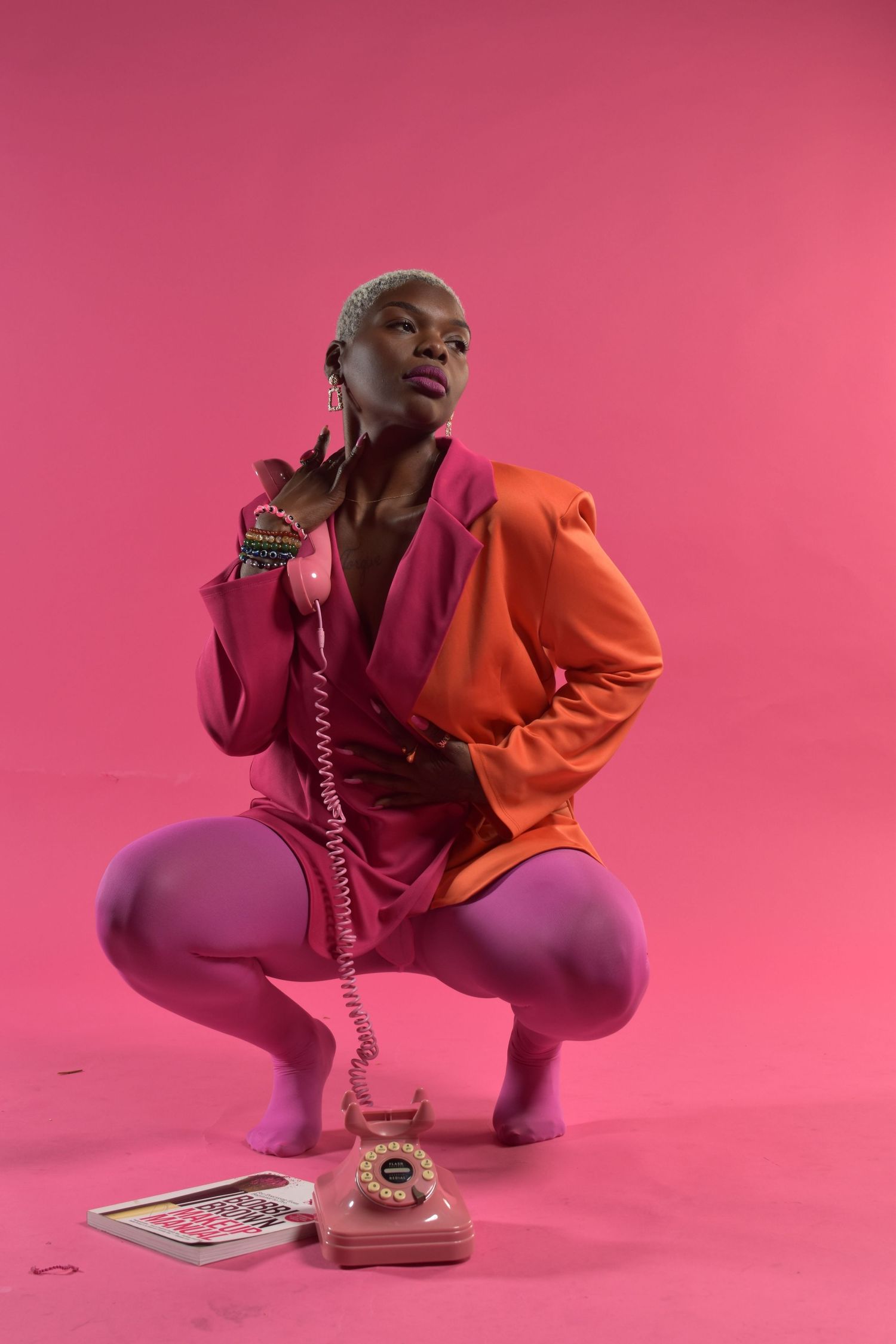 A Black woman in an orange and pink blazer crouches beside a vintage pink rotary phone against a pink background.
