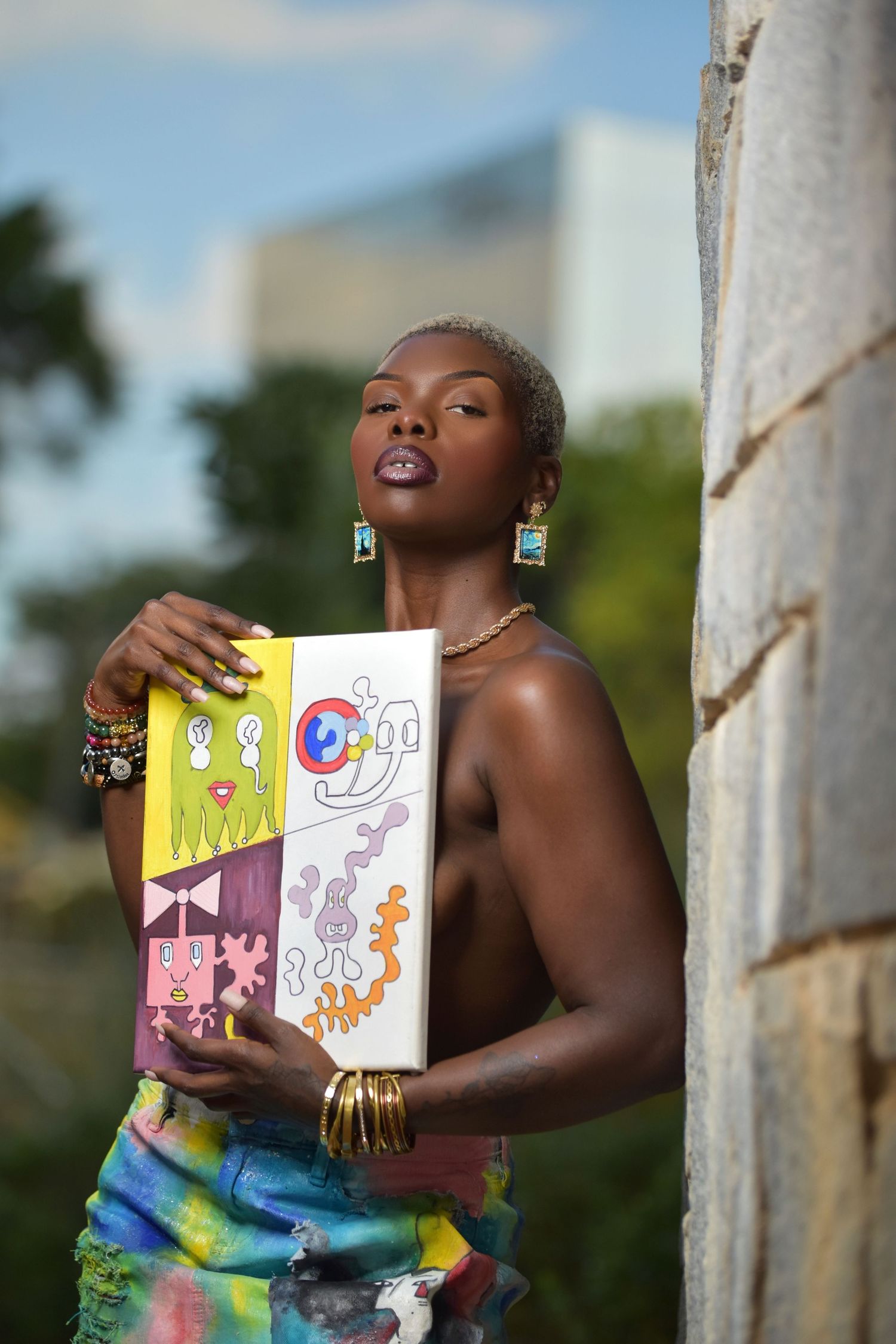 Black female artist holding vibrant illustrated canvas artwork outdoors, adorned with turquoise earrings and gold bracelets.