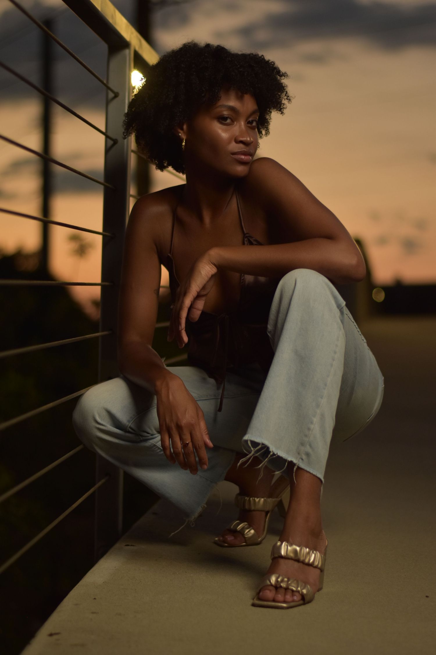 Black woman in brown crop top and light jeans crouching by metal fence during evening golden hour.