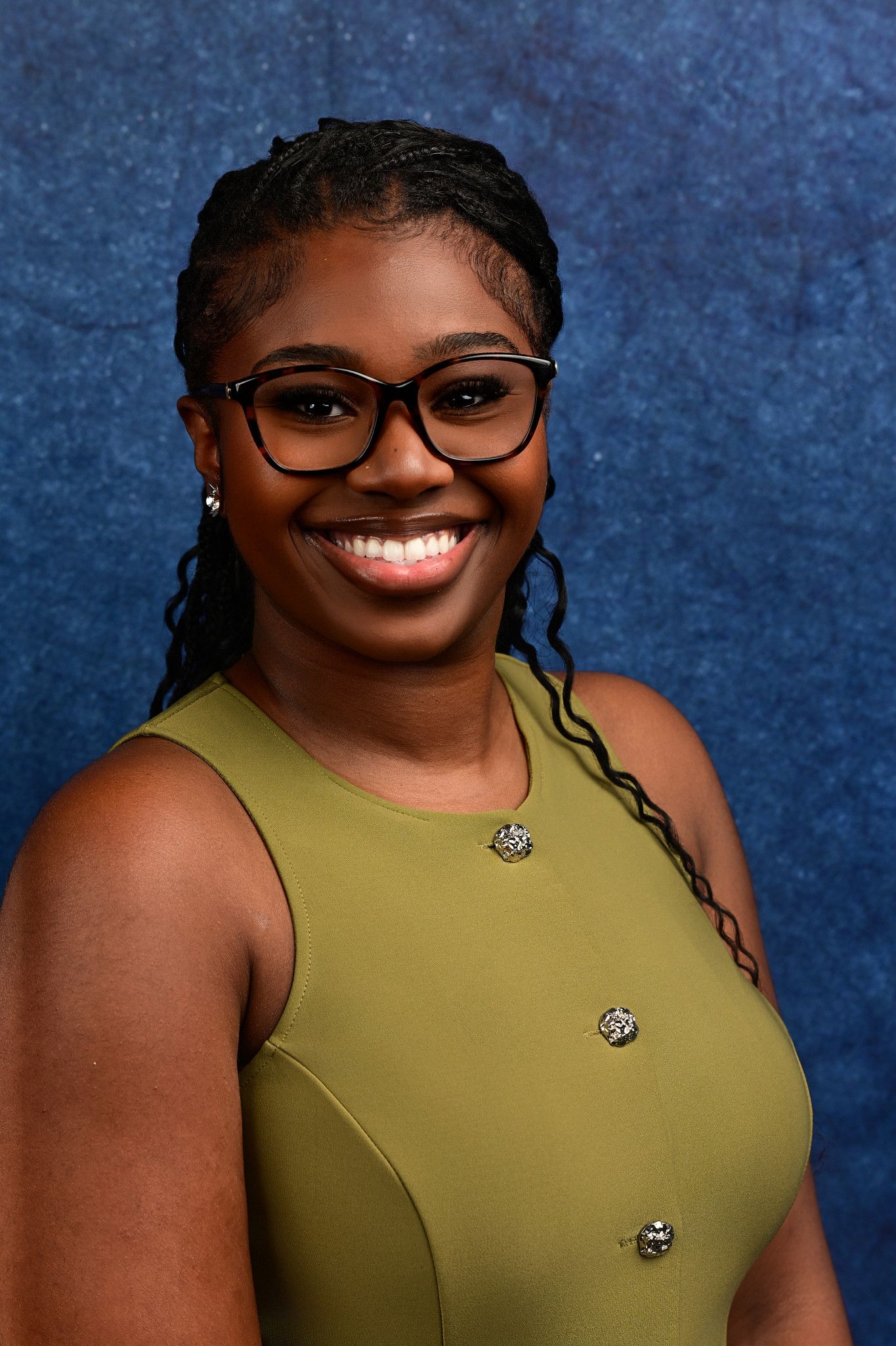 Smiling woman with long braids and black-framed glasses wears an olive green sleeveless top with decorative buttons against a blue backdrop.