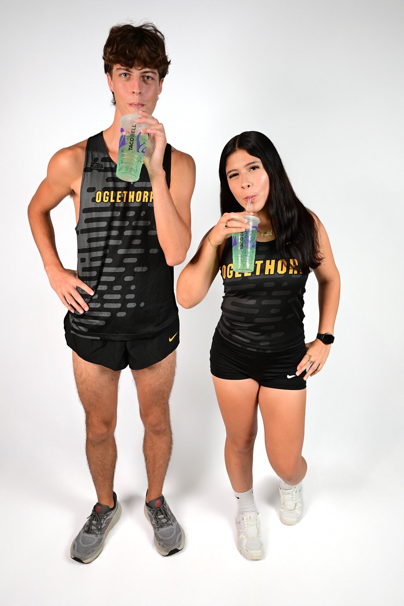 Two Oglethorpe athletes in black uniforms drinking from cups, posing on white background.