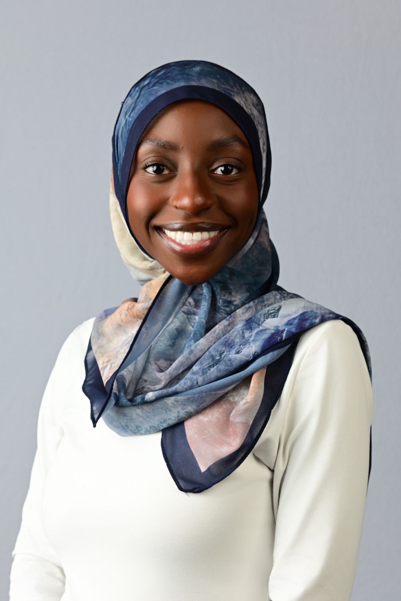 Smiling young Black woman wearing a blue floral hijab and white top, portrait photo on gray background.