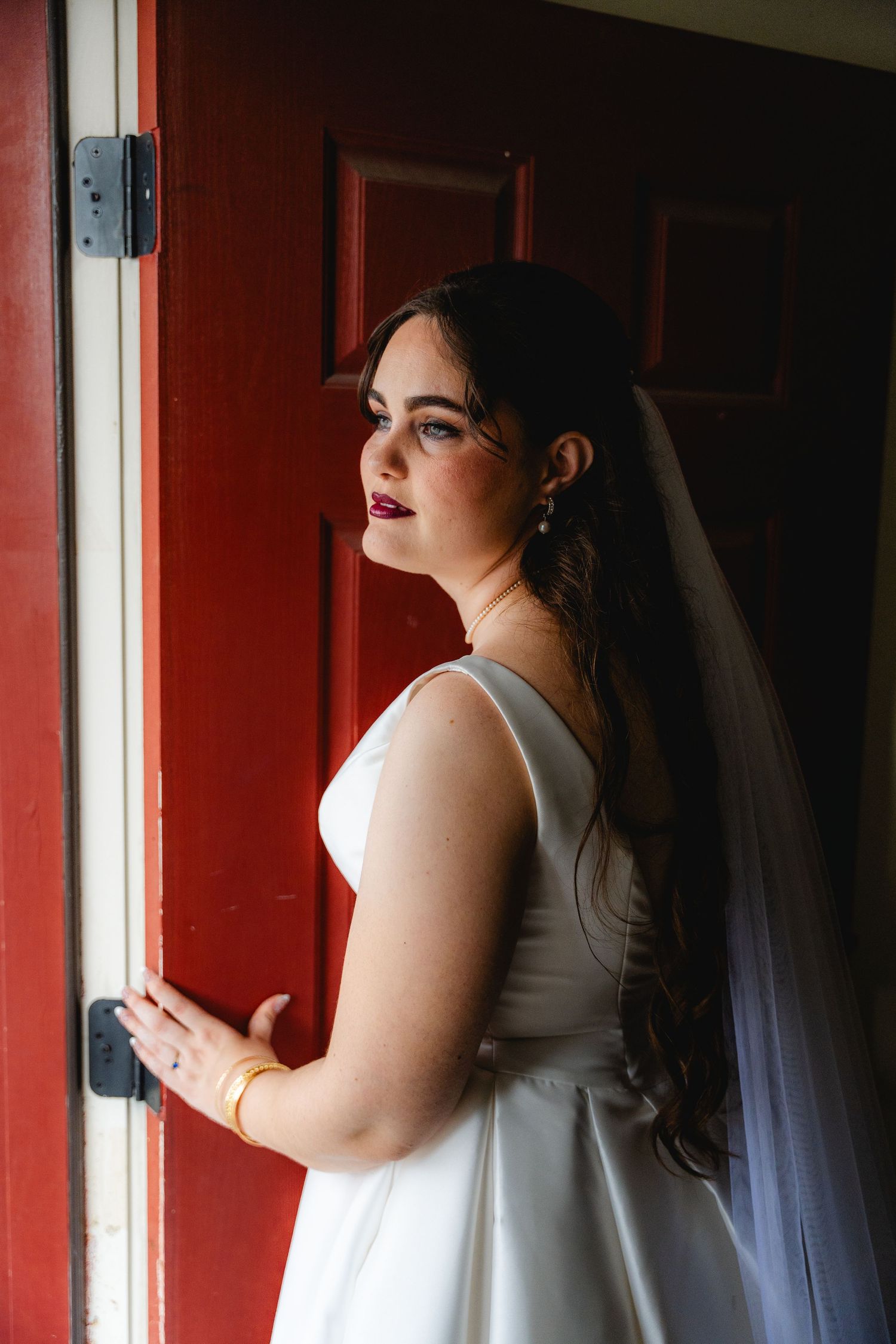 Bride in white gown and veil stands by a red door, gazing sideways with elegant makeup and gold jewelry in a Texas Wedding.