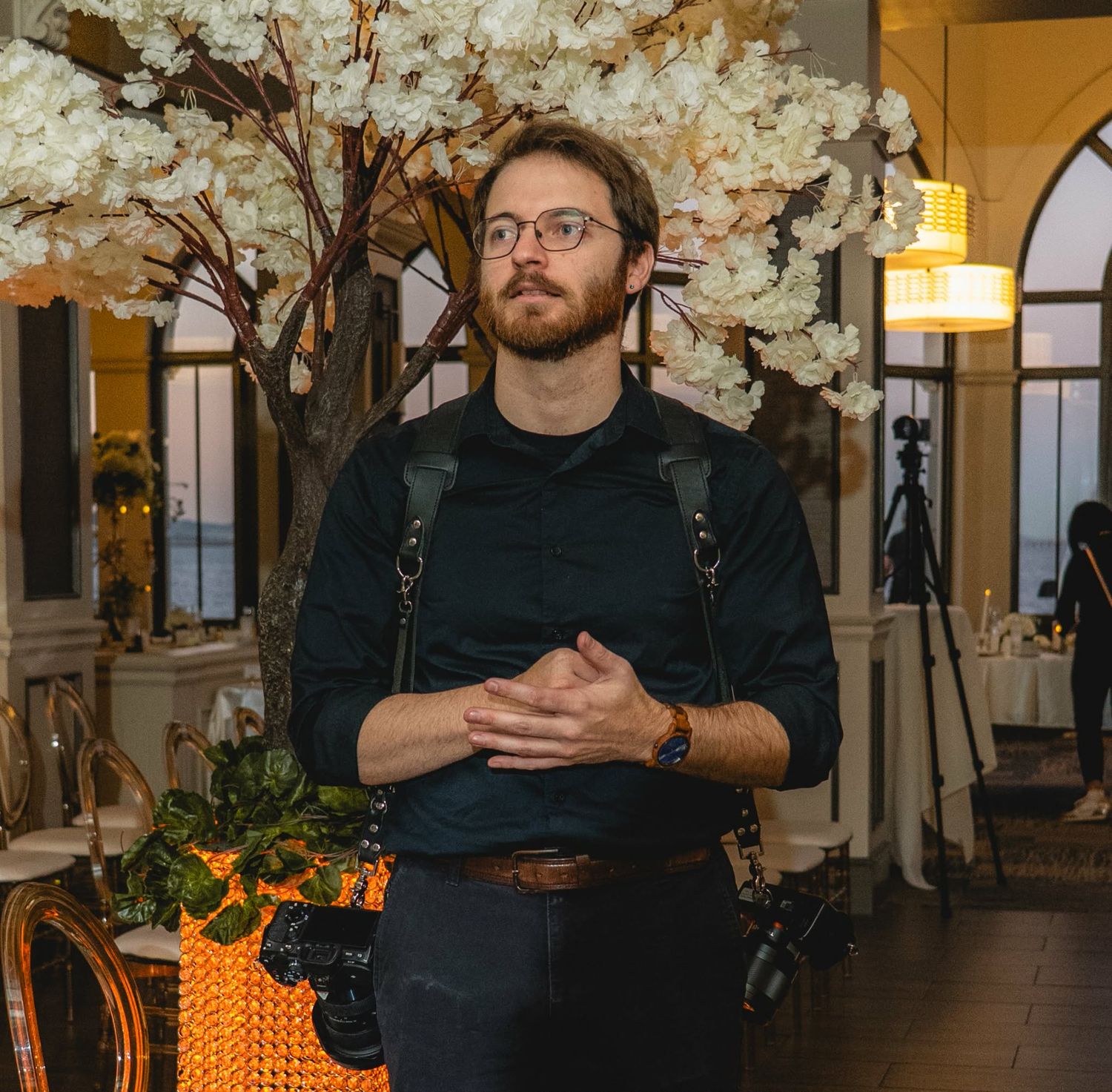 A bearded man with glasses wearing a black outfit and camera strap stands in an elegant floral decorated venue.