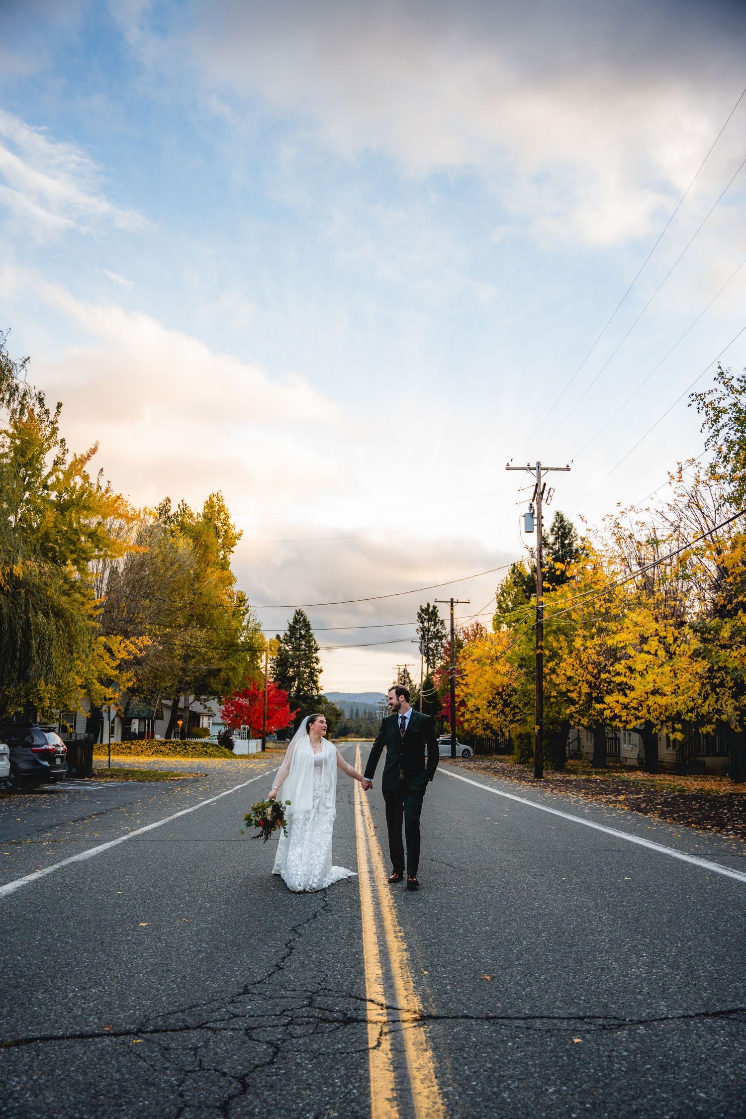 Bride and groom hold hands on a tree-lined road in autumn, wearing wedding attire under a blue sky at a Mt. Shasta wedding.