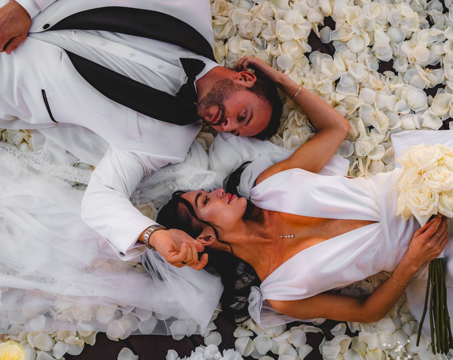 Bride and groom lying among white rose petals, aerial view of wedding couple in elegant attire with bridal bouquet.