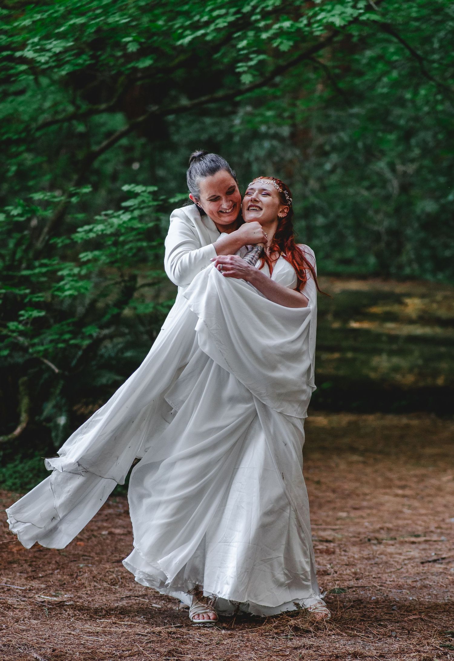 Two brides in white dancing joyfully on a forest path surrounded by lush green trees in a Jedediah state park.