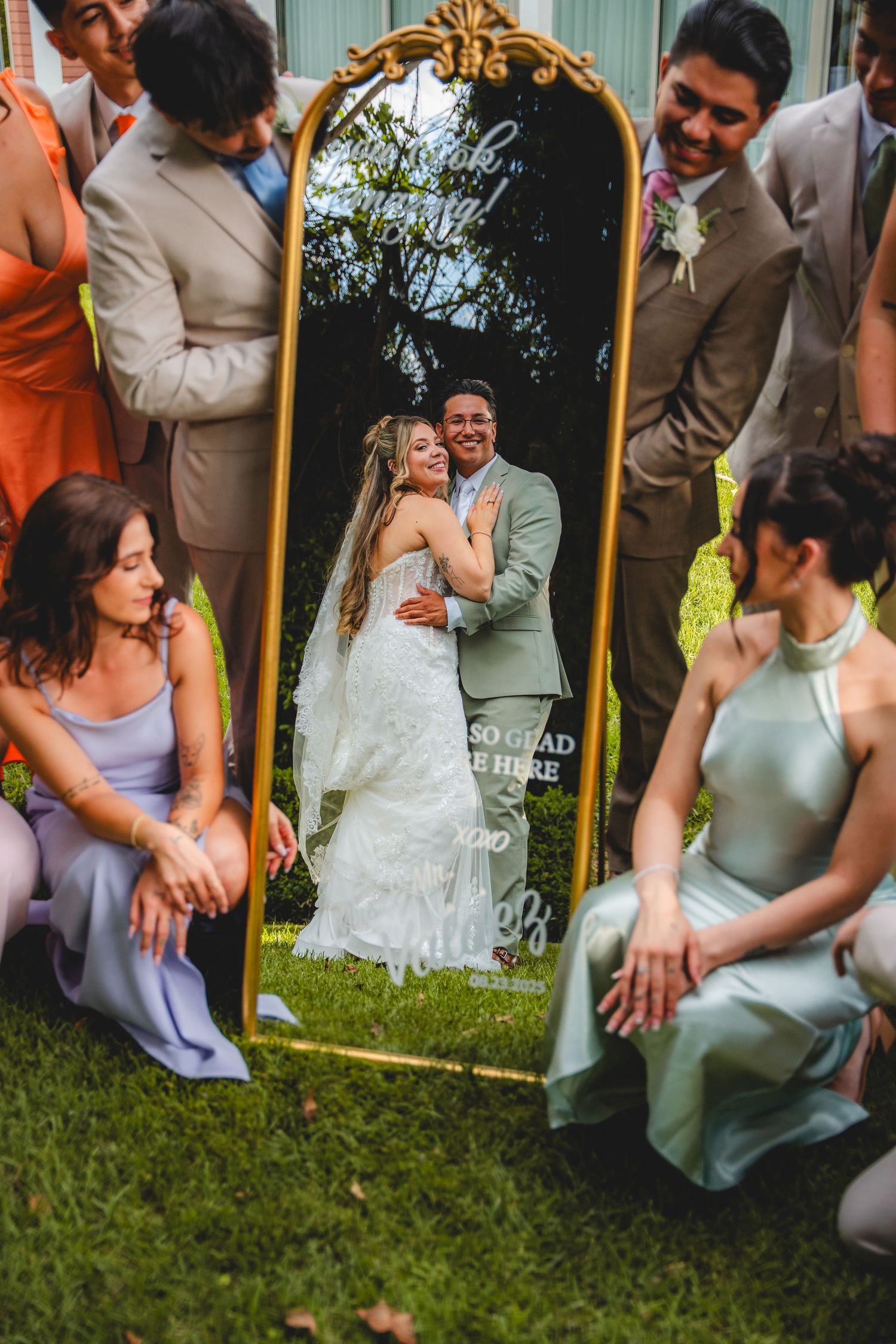 Bride and groom embrace reflected in ornate gold mirror surrounded by bridesmaids and groomsmen at outdoor wedding.