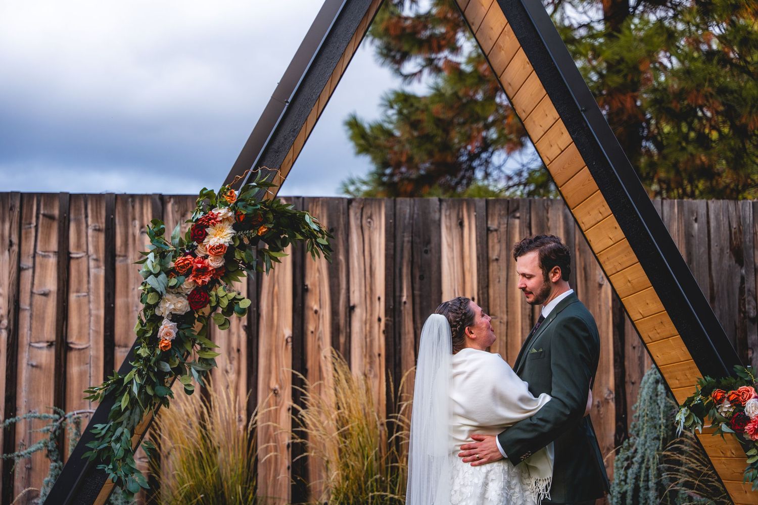Couple embraces at outdoor wedding ceremony under triangular arch adorned with red and green floral arrangements at the Shasta View Lodge.