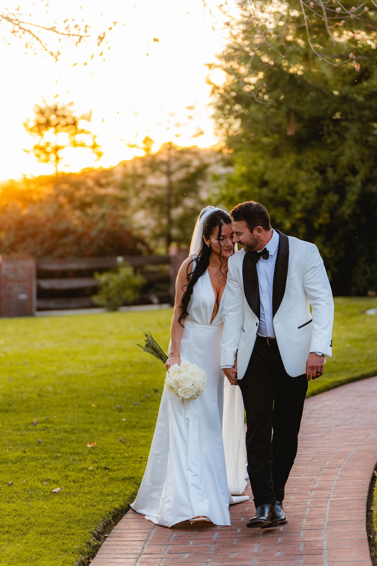 Bride and groom walking hand in hand on a brick path at sunset, bride holding white bouquet in elegant white gown.