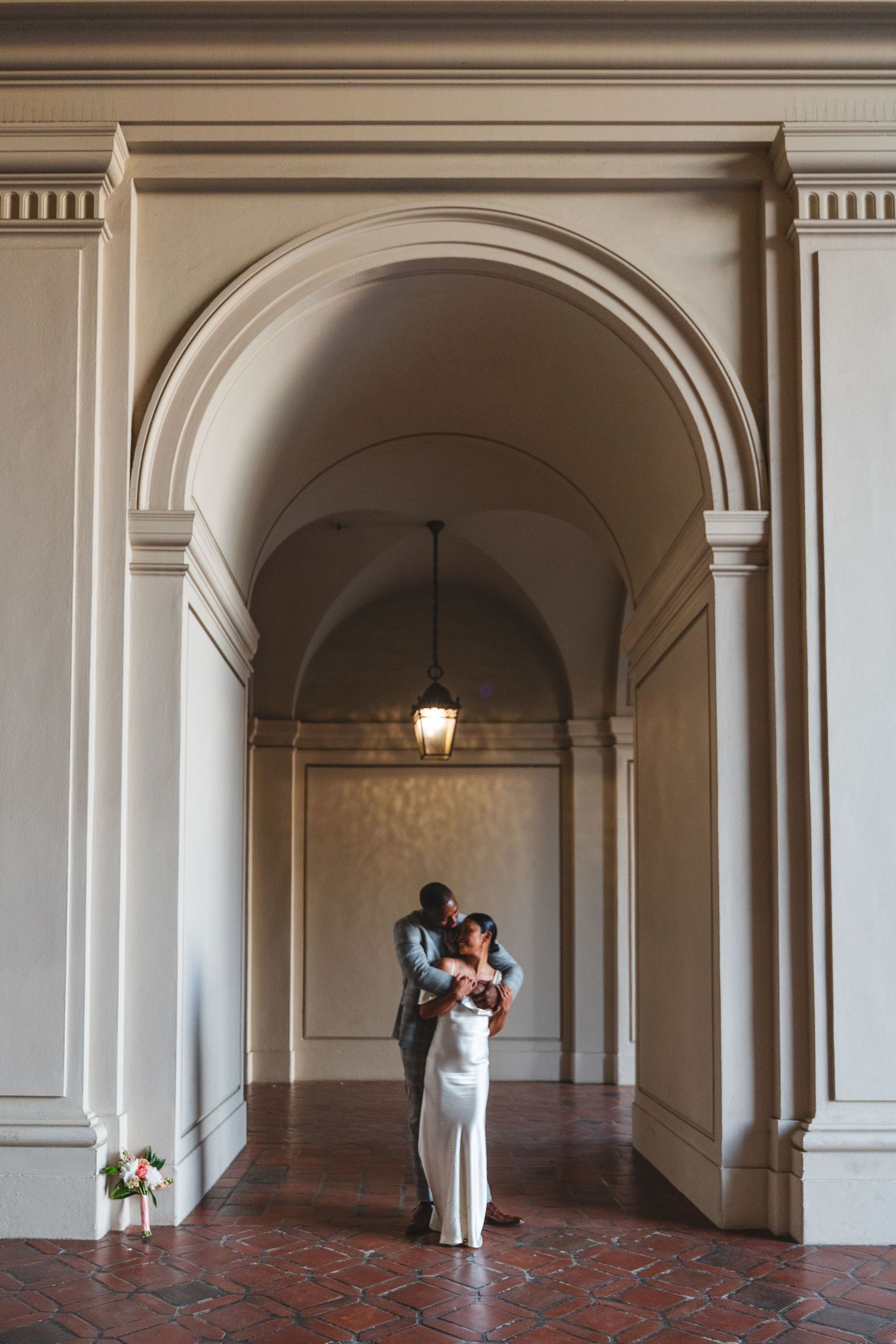 Couple embraces under elegant arched hallway with warm light, bride in white gown and groom in gray suit at a Pasadena City Hall Wedding.