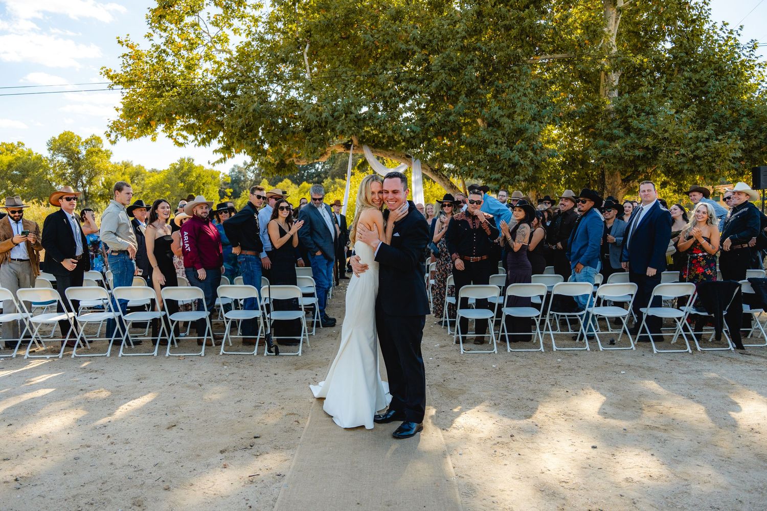 Bride and groom embrace at outdoor wedding ceremony with guests seated in white chairs under lush green trees.