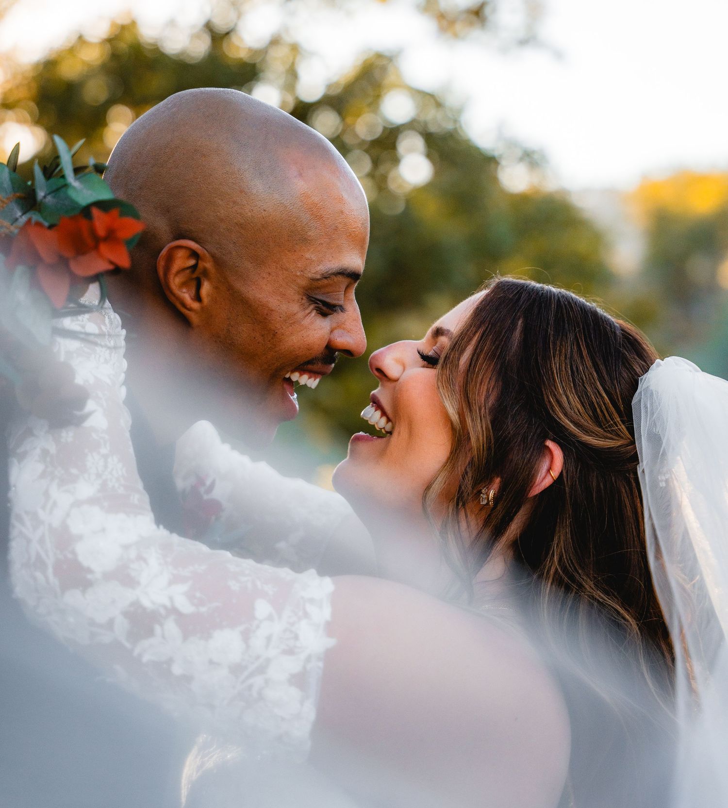 Joyful newlywed couple sharing a romantic moment, bride in lace gown with veil, groom with floral boutonniere outdoors.