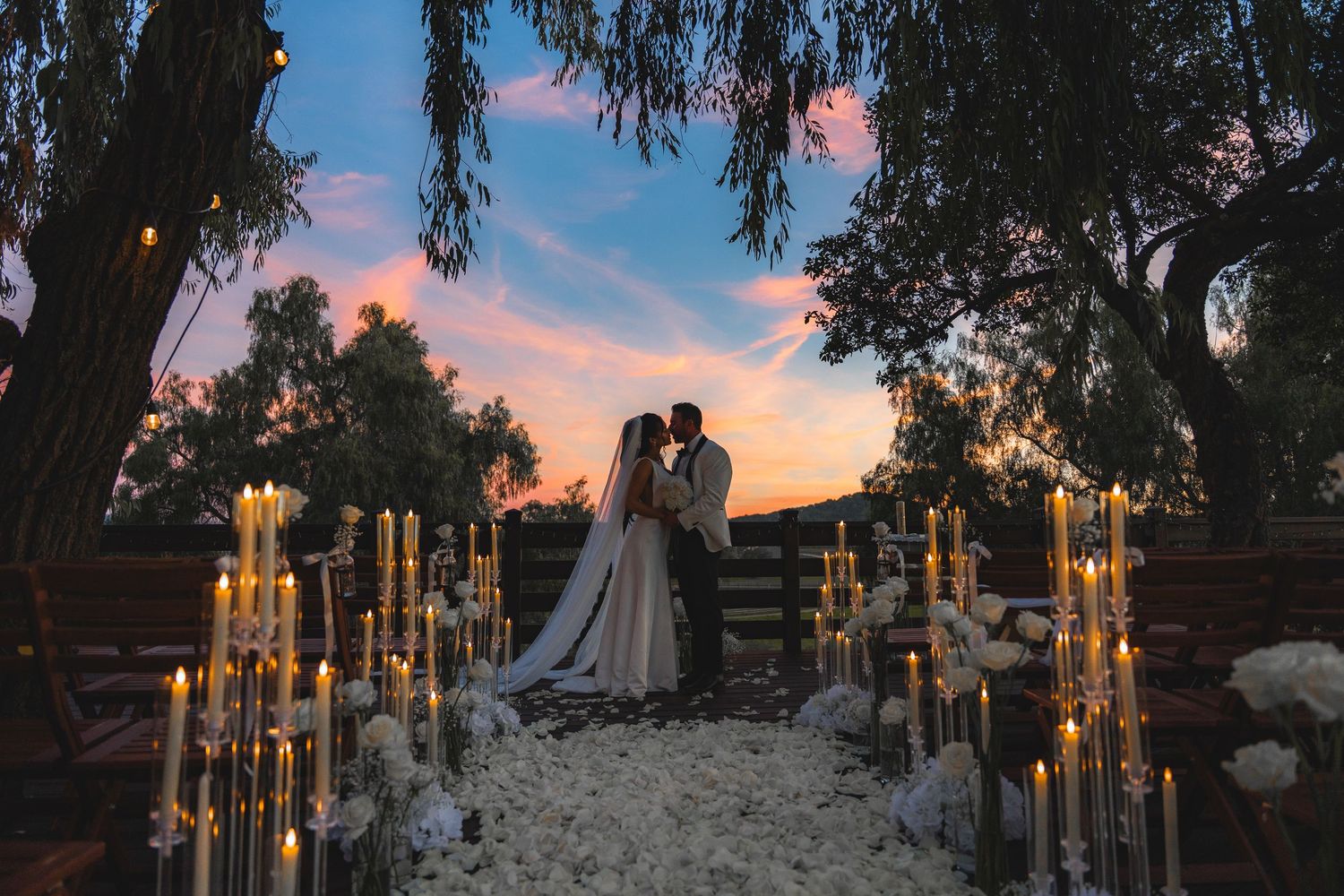 Romantic outdoor wedding ceremony at sunset with candles lining the aisle and couple kissing under trees