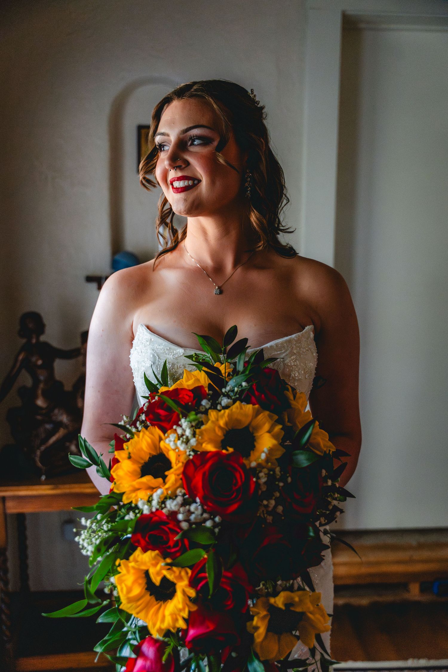Smiling bride in lace gown holding vibrant bouquet of sunflowers and red roses indoors.