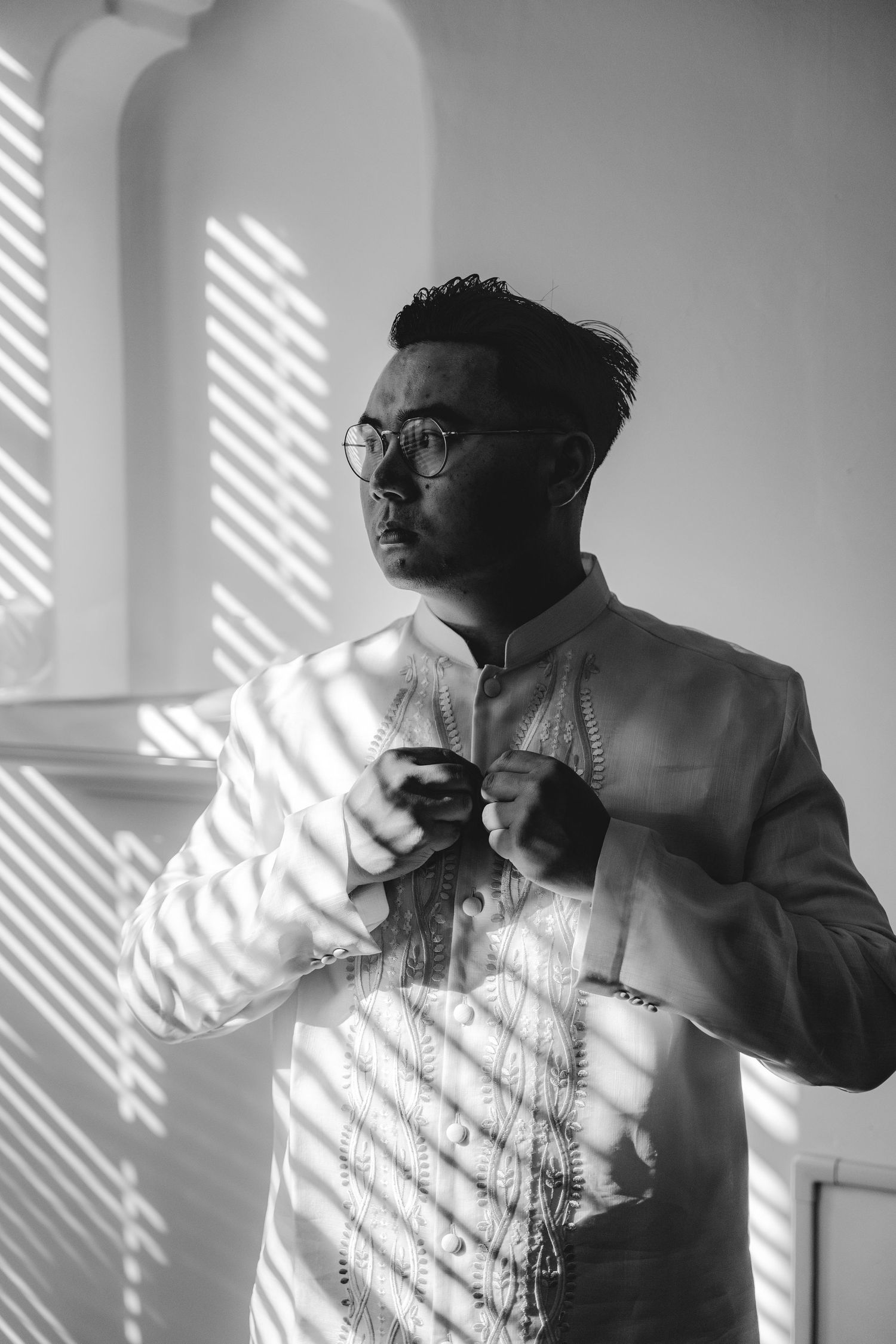 Black and white portrait of a man in embroidered wear adjusting his outfit near a window with dramatic light at a San Jose wedding