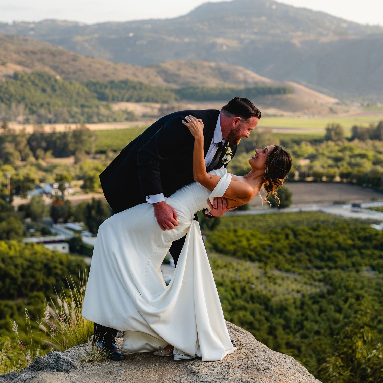 Bride and groom share romantic dip kiss on rocky hilltop overlooking scenic valley and mountains at golden hour wedding.