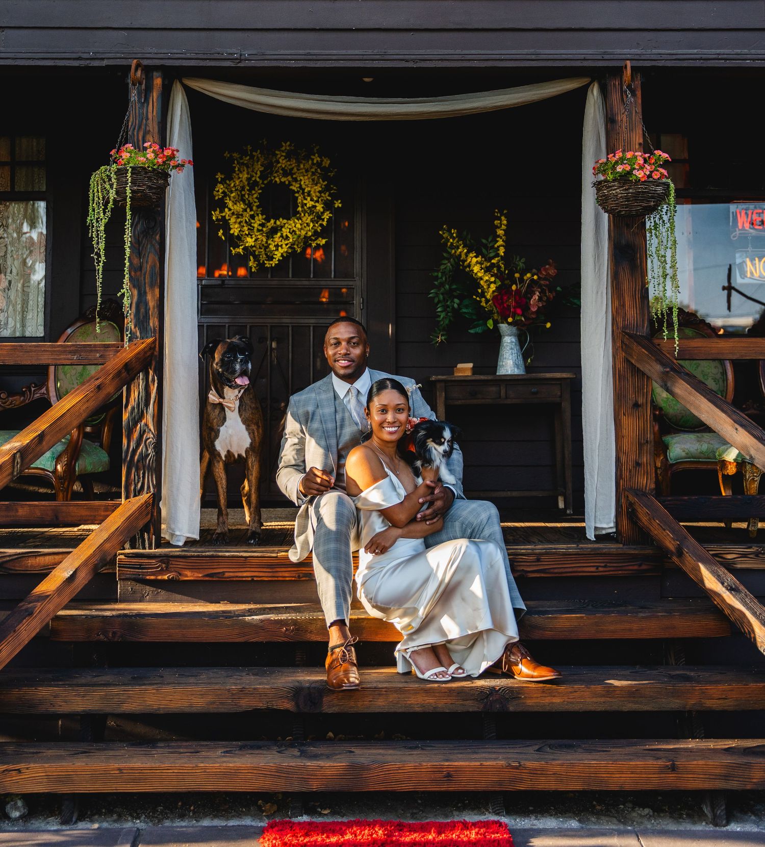 A couple in wedding attire sits on wooden steps with two dogs, surrounded by floral decor on a rustic porch at the Old Brown House venue.