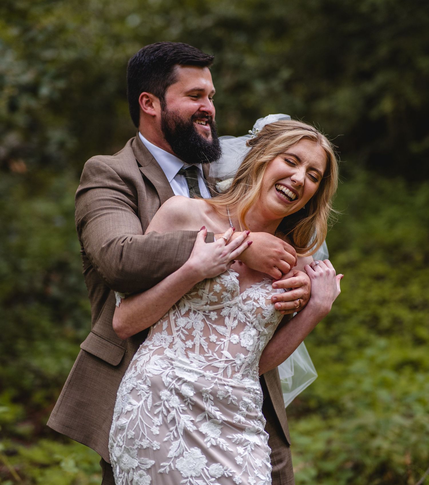 A laughing bride in lace gown embraced by bearded groom in brown suit outdoors an elopement in Big Sur, California