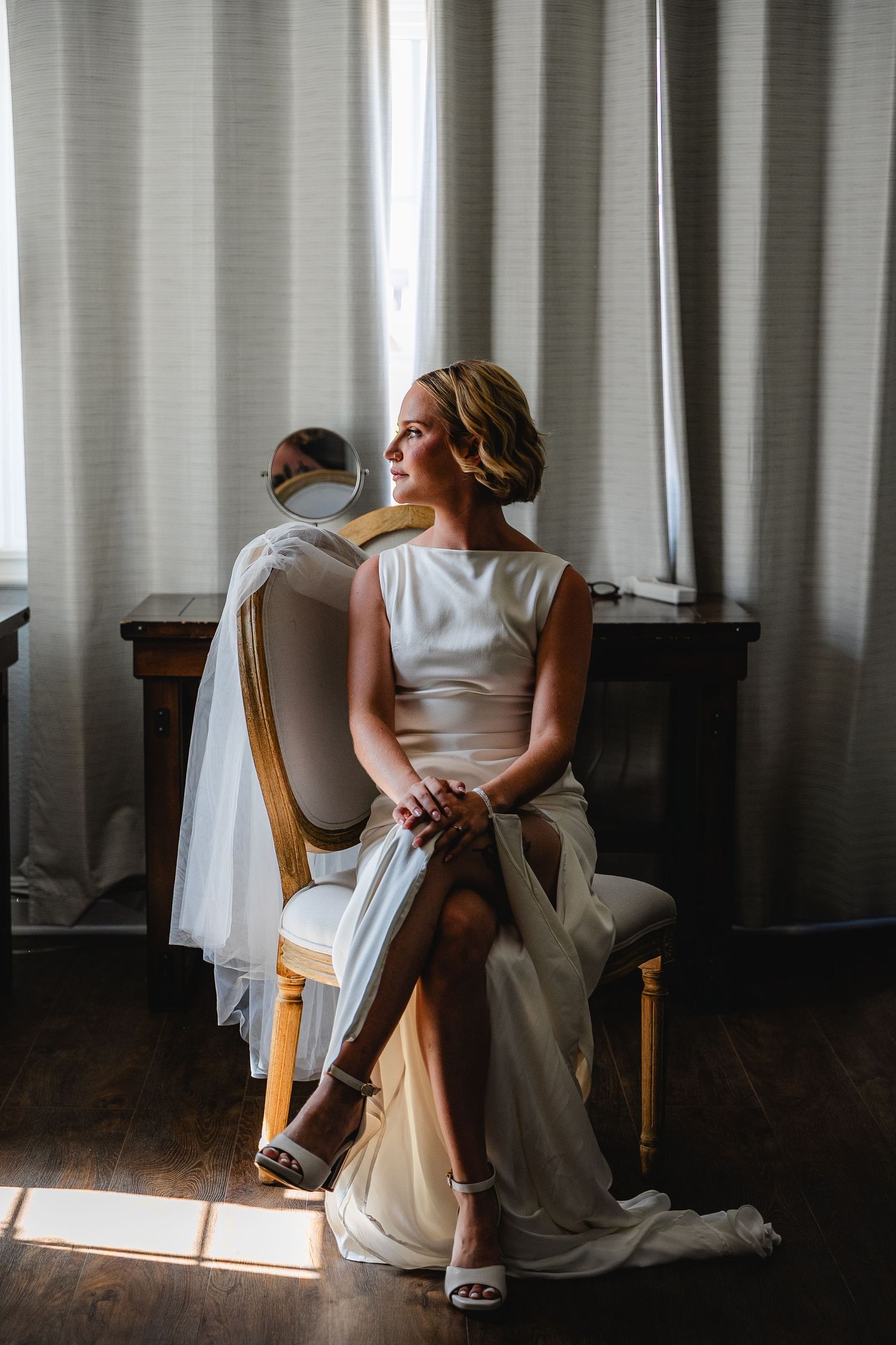 Bride in white gown sits gracefully by a sunlit window, reflecting in a mirror at a wooden vanity at a Casi Cielo wedding.