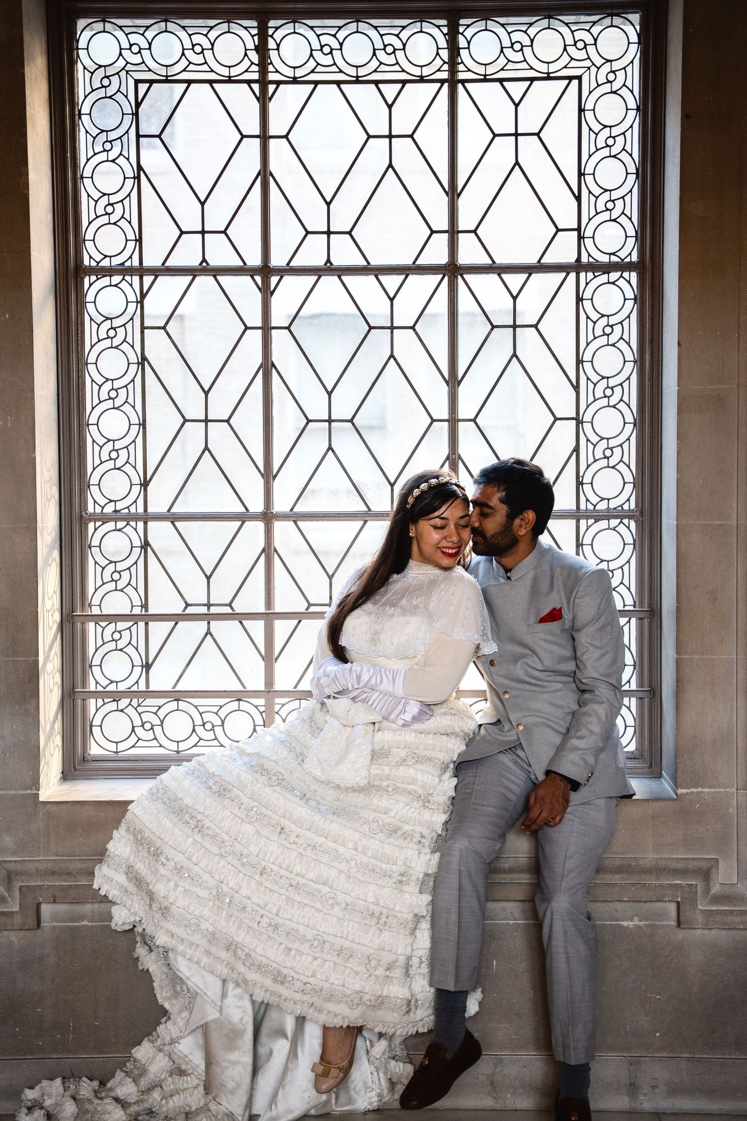 Bride in white ruffled gown and groom in gray suit share a romantic moment by an ornate stained glass window at the San Francisco City Hall.