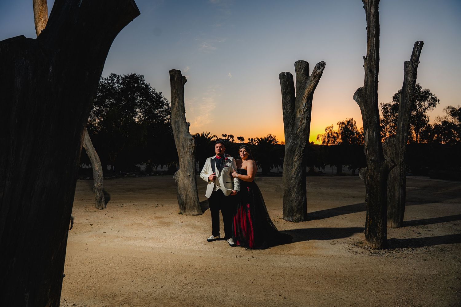 Couple embracing at sunset among bare trees in a desert landscape, woman in red gown, man in white suit at a Galway Downs wedding.