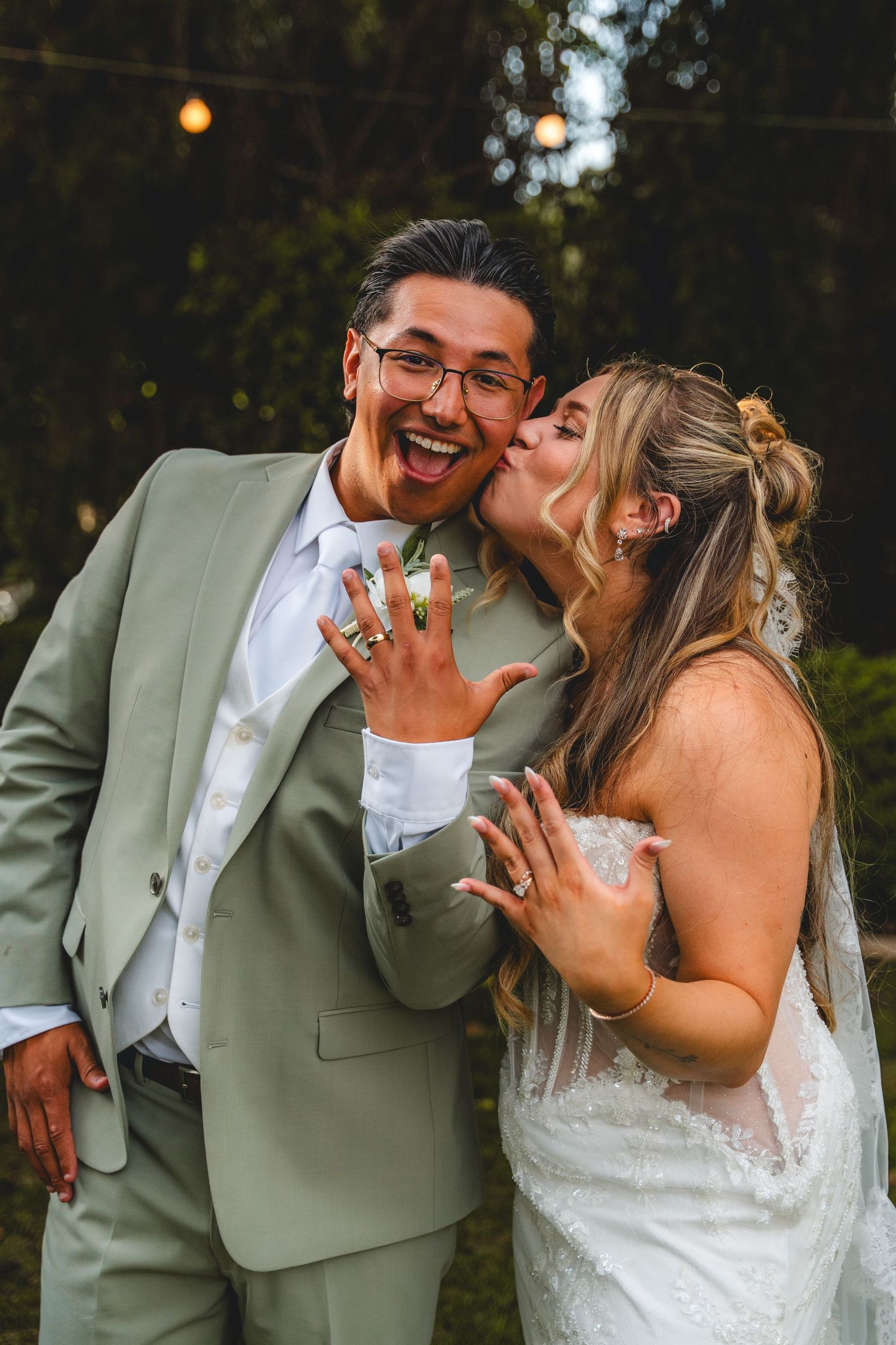 Joyful newlyweds showing wedding rings, bride in lace gown kissing groom in green suit at a San Diego Wedding at The Handlery Hotel
