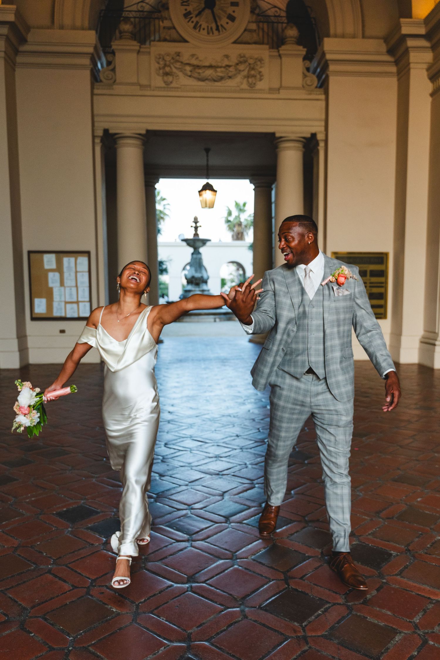 Joyful bride in white gown and groom in plaid suit dancing and laughing in elegant venue hallway in Pasadena City Hall