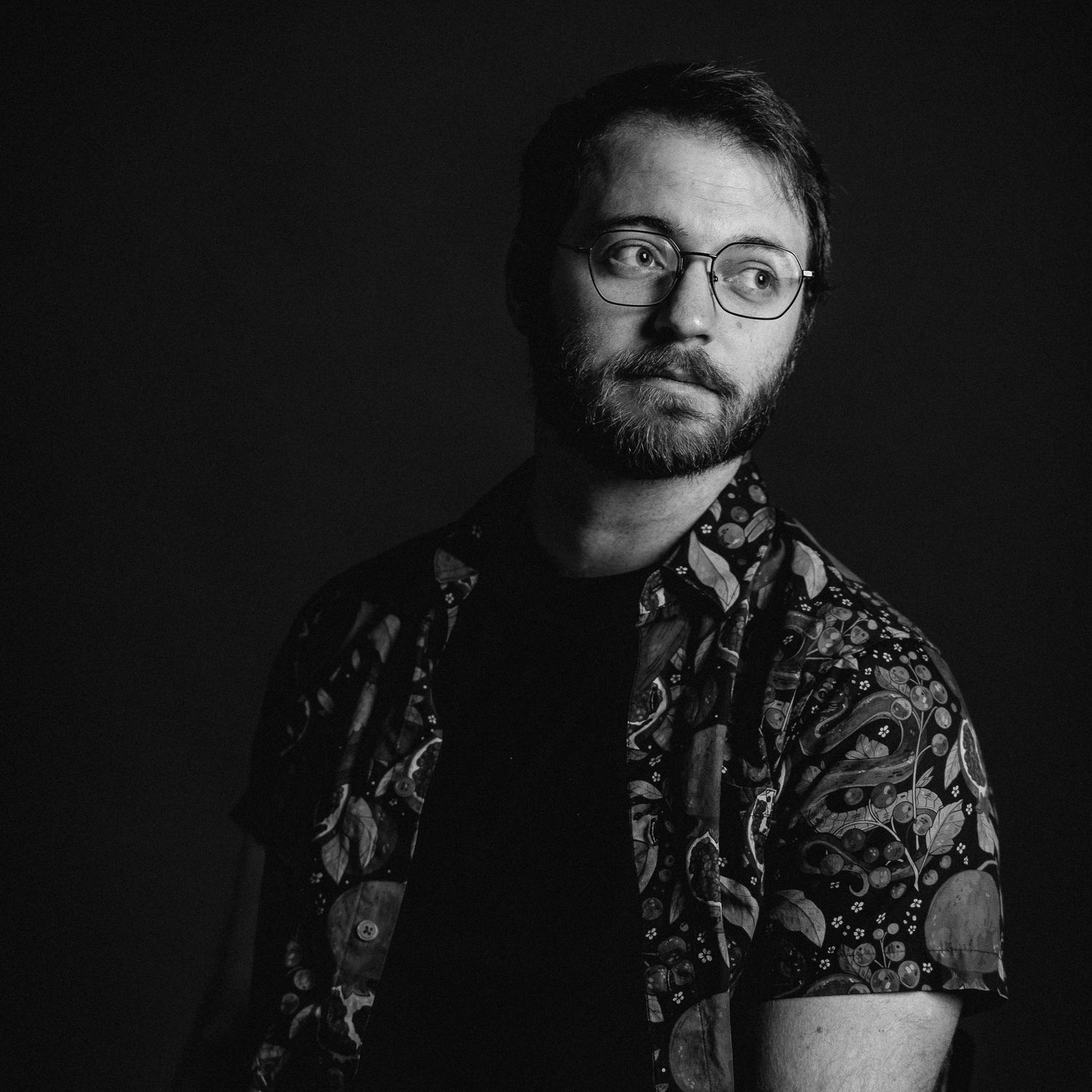 Black and white portrait of a young bearded man wearing glasses and a floral shirt against a dark background.