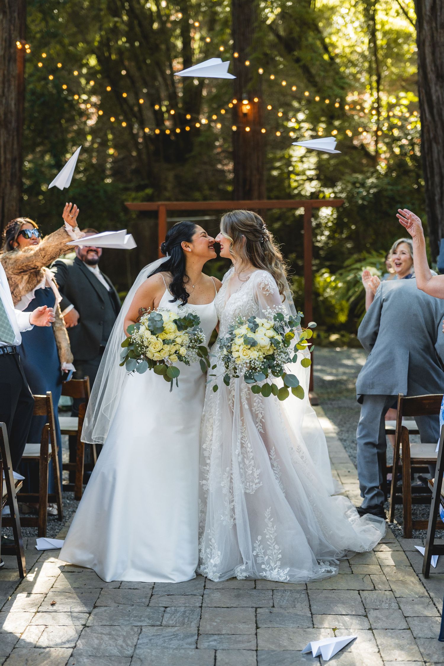 Two brides in white gowns kiss at outdoor wedding ceremony while guests toss paper airplanes in a redwoods venue.