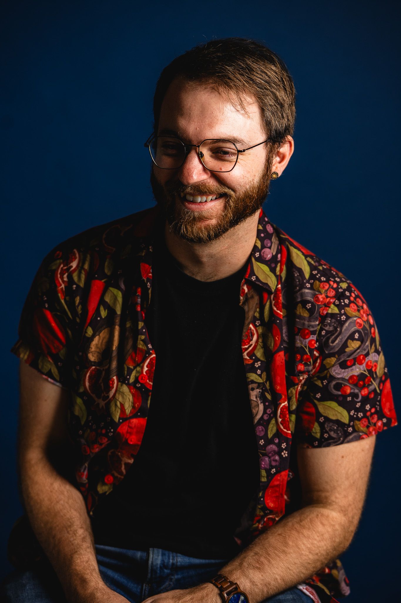 Smiling bearded man with glasses wearing a colorful floral shirt over a black tee, posed against a dark blue background.