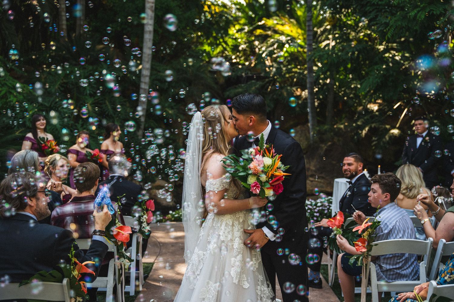 Bride and groom share first kiss surrounded by bubbles at a Grand Tradition wedding ceremony with seated guests.
