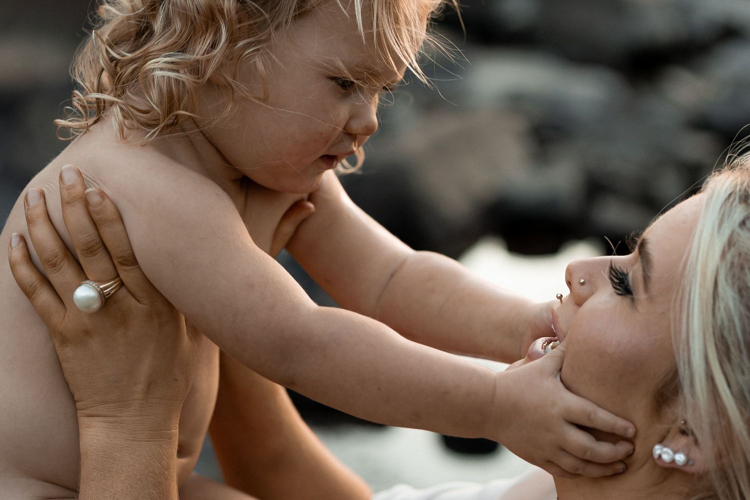A mother lovingly holds her toddler close, both sharing a tender moment outdoors near rocky shores.