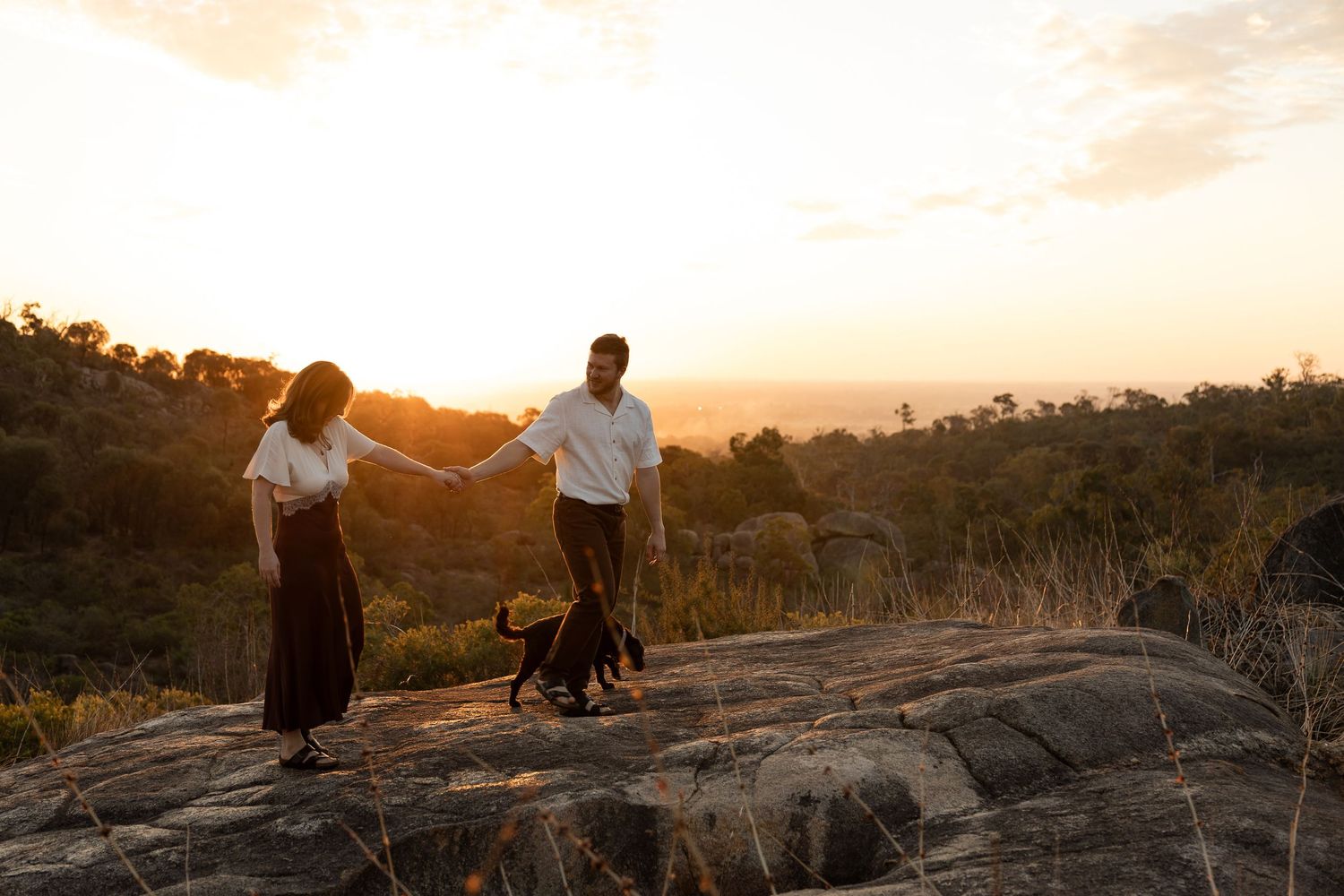 Couple holding hands with their black dog at sunset on rocky hilltop overlooking lush green landscape of the Perth hills