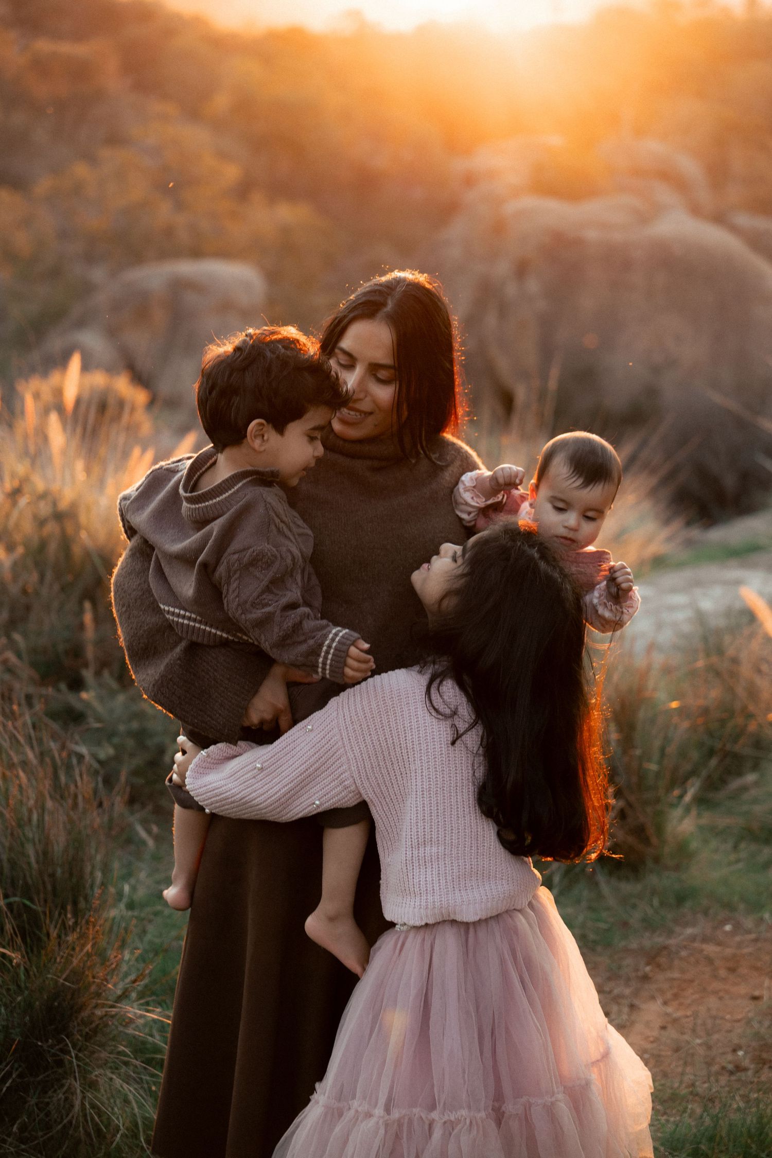 A mother holds two young children at golden hour outdoors, while her daughter in a pink skirt hugs her warmly.